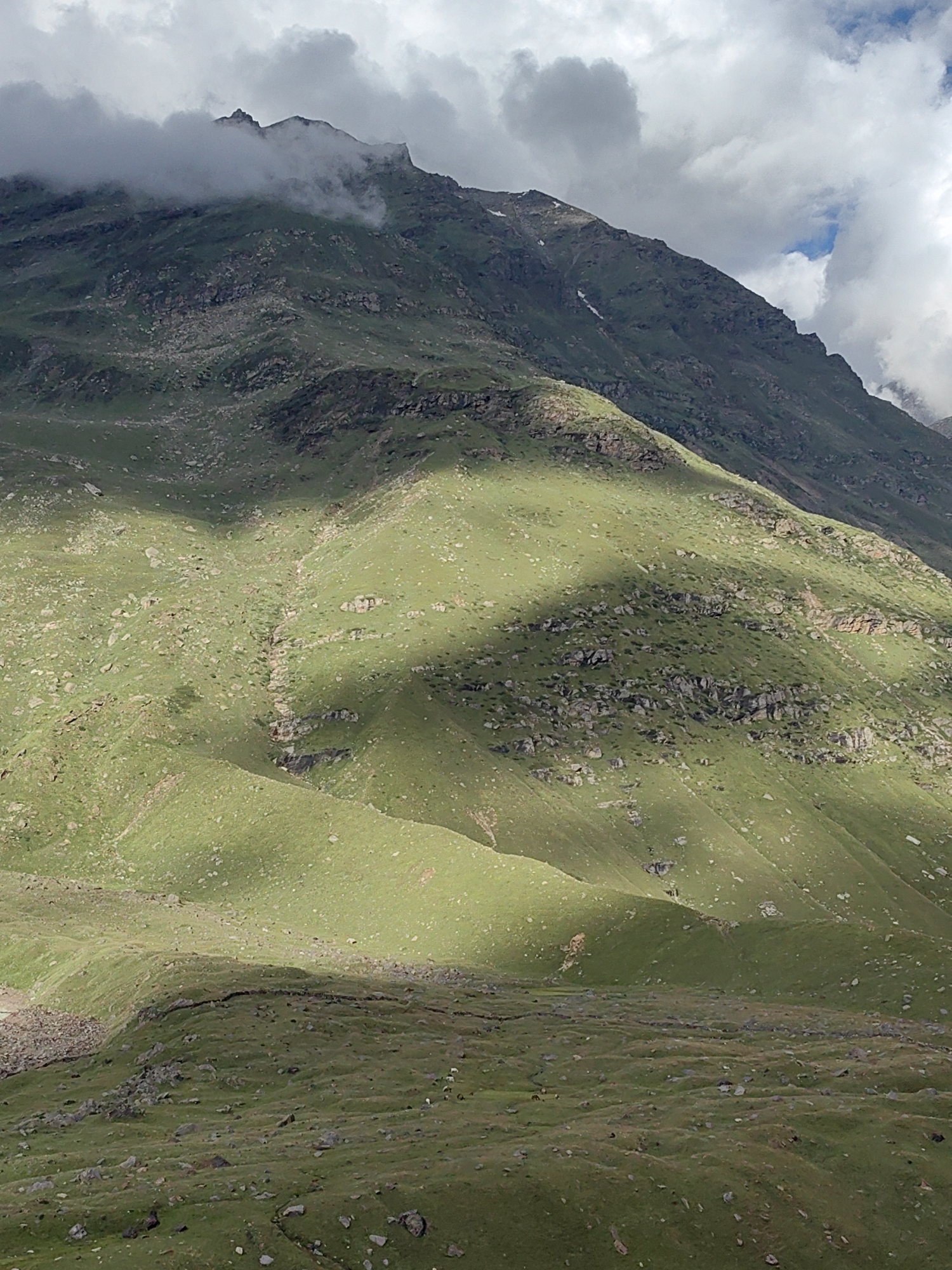 The image shows a mountainous landscape in Burwa, India. The mountains are covered in green grass and dotted with rocks. The sky is partly cloudy, with some of the clouds obscuring the peaks of the mountains. Sunlight is casting shadows across the mountainside, creating a dramatic contrast between light and dark areas. There are no people visible in the image. The environment is natural and wild, with no signs of human development other than what might be inferred by the location being a city.