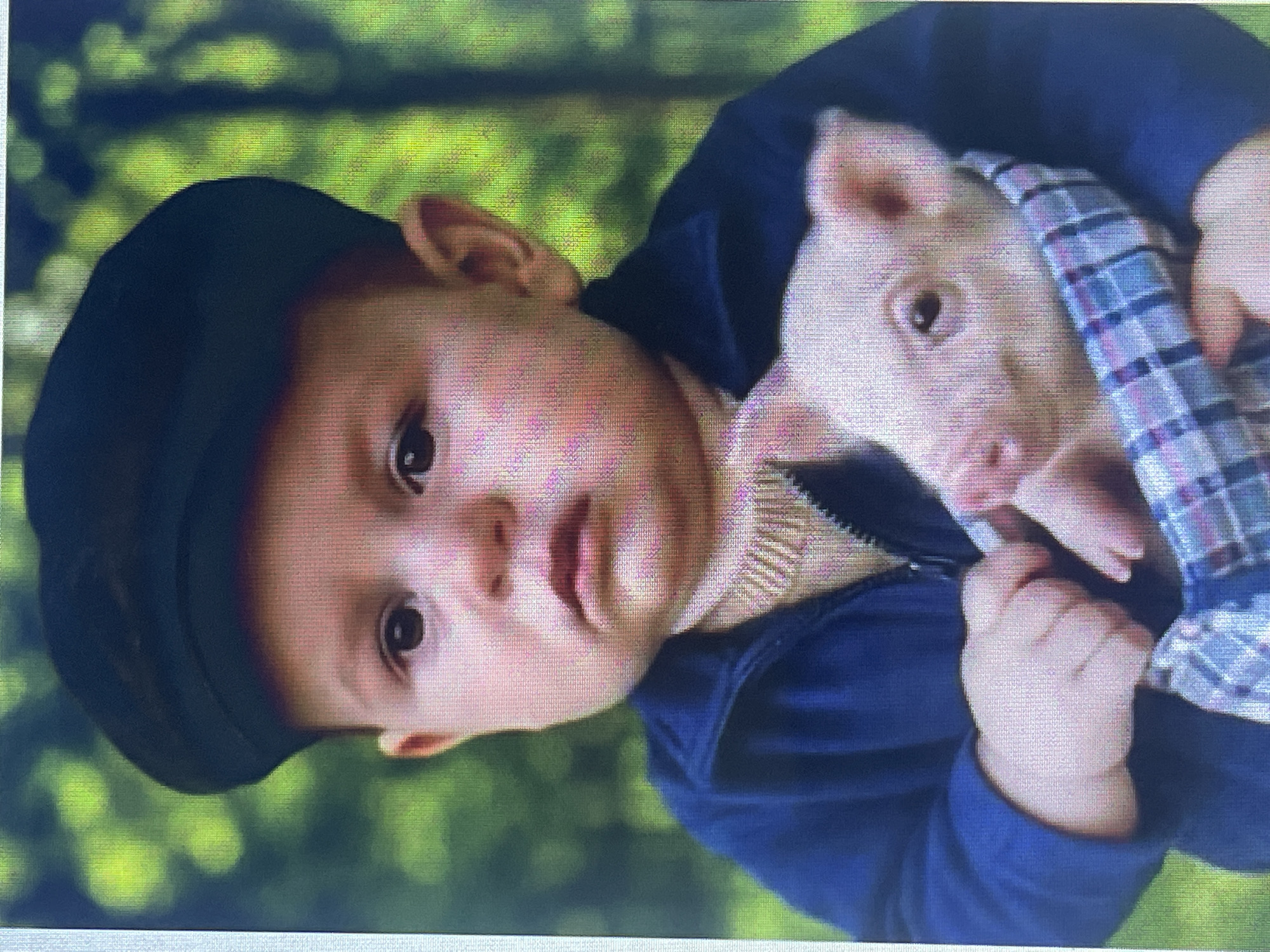 A young child, appearing to be a toddler, is shown wearing a dark cap and a navy blue zippered jacket over a cream-colored sweater. The child is holding a small pink piglet, which is partially wrapped in a blue and white plaid cloth. The child's expression is somewhat somber. The background consists of blurred green foliage, suggesting an outdoor setting. The image's quality is pixelated, making precise identification of the location in Stary Oskol, Russia impossible.