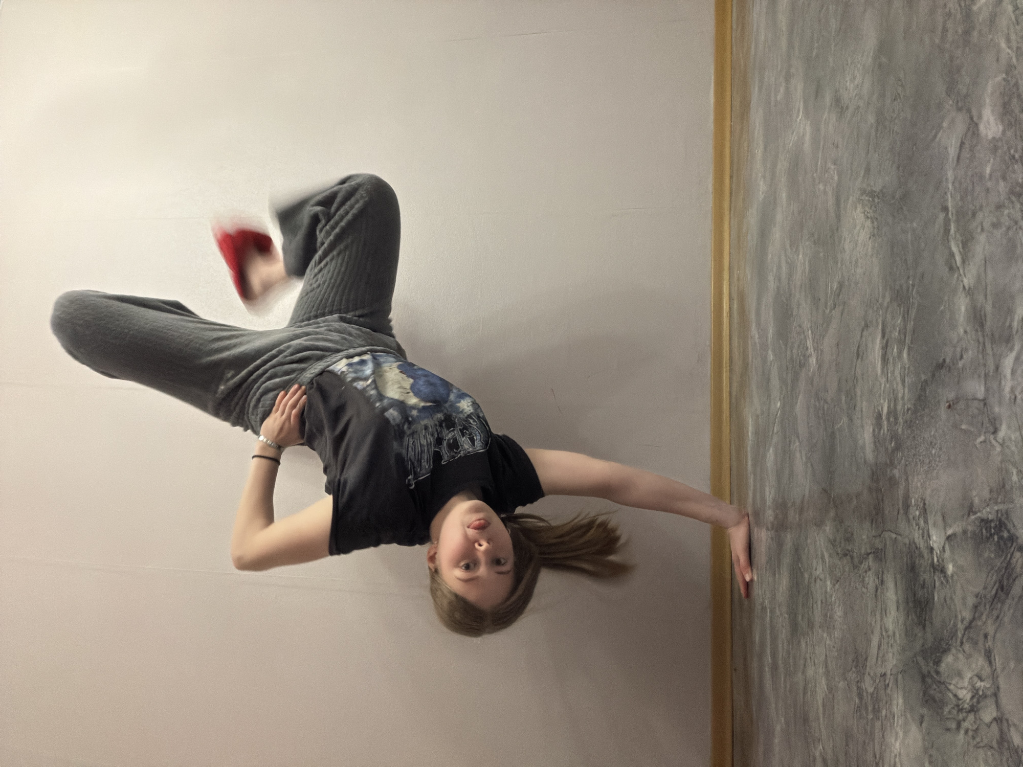A young individual, appearing to be female, is depicted in an indoor setting, posing in an inverted and relaxed position. The person is lying on their back on a plain, light off-white surface, which appears to be the floor. Their legs are bent at the knees and elevated, with feet pointing upwards. One foot wears a distinct red shoe, which shows slight motion blur, while the other leg and foot are partially obscured.
The subject wears a black t-shirt with a blue, white, and grey graphic print, and baggy grey sweatpants. Their light brown hair is pulled back in a ponytail. Their head is tilted back, looking directly at the camera, with a playful expression, including the tongue slightly out. One arm is bent across their torso, while the other arm is extended to the side, hand pressing against a dark grey, textured wall. This wall has a marbled or concrete-like finish and is separated from the plain light wall (which could be another wall or the ceiling, given the inverted pose) by a thin, golden-brown trim. No visible text is present. The overall impression is a casual, dynamic, and somewhat playful moment captured indoors.