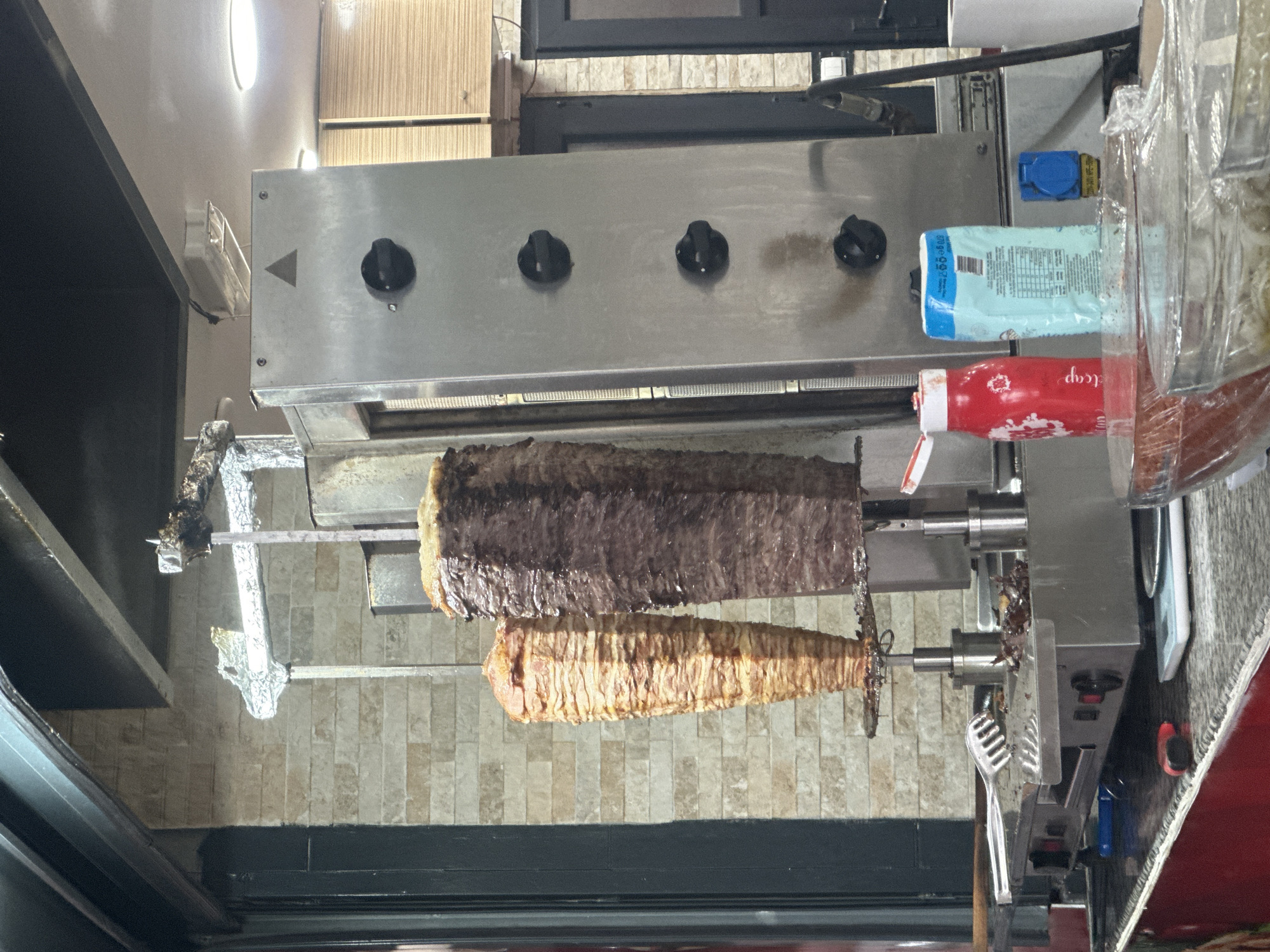 A shawarma or doner kebab cooking apparatus is presented vertically. Two stacks of meat are impaled on vertical skewers and heated by a gas-powered grill above. The top meat is dark brown, whereas the bottom is a paler tan. A ketchup bottle sits to the right of the grill, and a package of napkins is visible. The backdrop consists of off-white, rectangular tiling. The grill has four knobs that may regulate the heat, as well as a triangular sticker towards the corner of the grill. This image was most likely taken in Fethiye, Turkey.