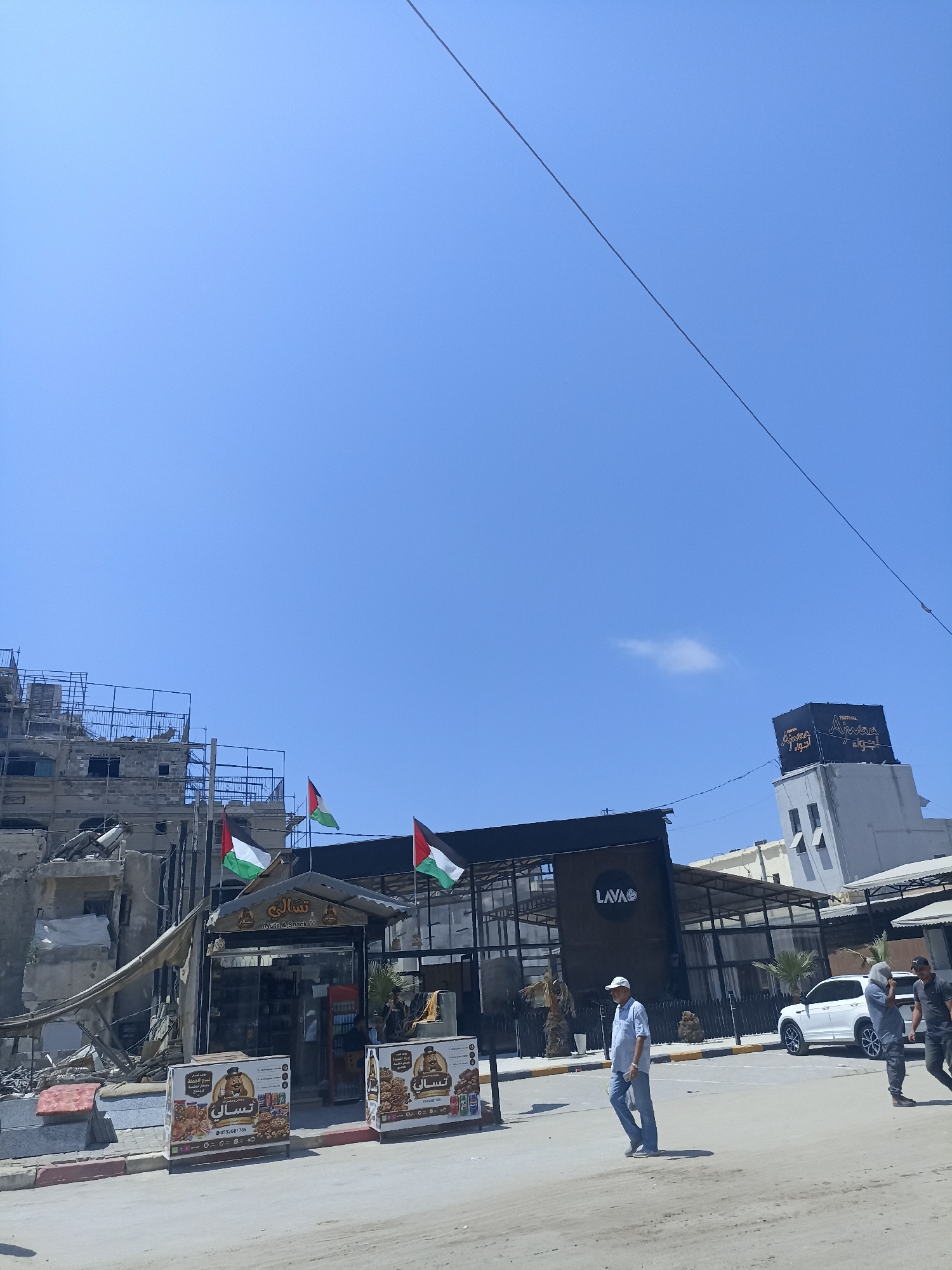 In the city of Gaza, Palestinian Territories, a street scene unfolds under a clear blue sky with a single wire diagonally bisecting the upper portion of the frame. The foreground features a dusty road with a curb. To the left, damaged buildings with exposed concrete and metal structures suggest recent destruction. A tarp is draped over some of the debris. In front of these ruins, two large advertising boards display images of food items and Arabic text.
To the right of the damaged buildings is a small shop with a peaked awning. Two Palestinian flags are prominently displayed above the shop entrance, which features a sign that reads "Nuts & Snack" in English and Arabic. The shop appears open, with products visible inside.
Further to the right, a modern building with dark windows and a black awning is visible. A circular logo reading "LAVA" is displayed on this structure. A white car is parked to the right of this building, and three individuals are walking on the street in front of it. One man, wearing a blue shirt and jeans, walks towards the viewer's left. Another person, wearing a grey headscarf and dark clothing, walks in the same direction. A third person, in a dark shirt and pants, walks towards the viewer's right, slightly behind the other two. The environment appears to be an urban setting with a mix of damaged and intact structures.