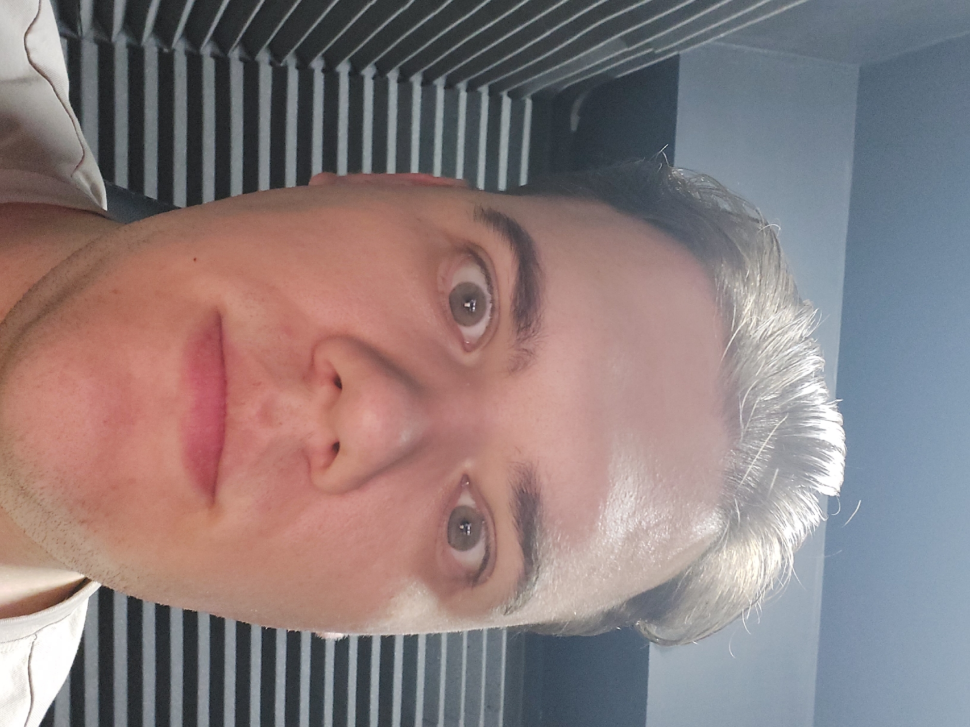A close-up, slightly off-center portrait shows a Caucasian male with light complexion and short, light brown hair. He has brown eyes, a prominent nose, and a slight stubble on his face. He appears to be indoors, with a striped black and white wall in the background. There's a plain, light-colored wall to his right. The image lacks specific details to identify the location within Tashkent, Uzbekistan, or determine any specific actions, interactions, or subjects present in the wider environment.