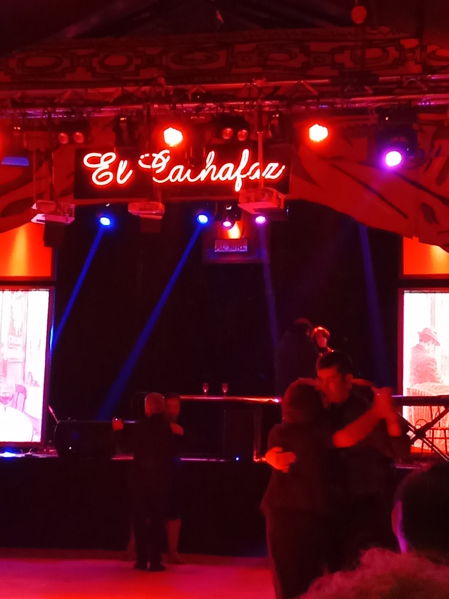 The image depicts a neon sign reading "El Bailecito" in a dimly lit venue, likely a dance hall or nightclub. Red and blue spotlights illuminate the space. In the foreground, a couple is captured mid-dance, appearing to be engaged in tango or a similar partner dance. The man is wearing a dark suit, and the woman is in a dark dress. Several other figures are visible in the background, some standing and others appearing to be seated. To the left, a large screen displays a black and white image, possibly a projection. The overall ambiance is one of performance and social gathering, with a focus on dance. The location is identified as Providencia, Chile, a commune known for its vibrant nightlife and cultural venues.