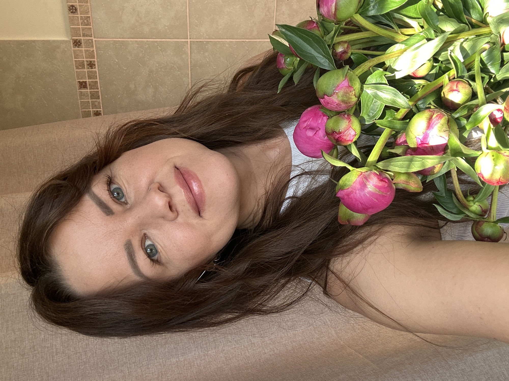 A mid-shot portrait of a woman in Chaikovsky, Russia, holding a bouquet of pink peonies. The woman has long, dark brown hair, blue-green eyes, and is wearing a white top. She is smiling gently and looking at the camera. The background consists of a light beige curtain on the left and a tiled wall with a patterned border on the right. The sunlight highlights the flowers and the woman's face. The scene appears to be indoors, possibly in a bathroom or dressing area given the tiling.