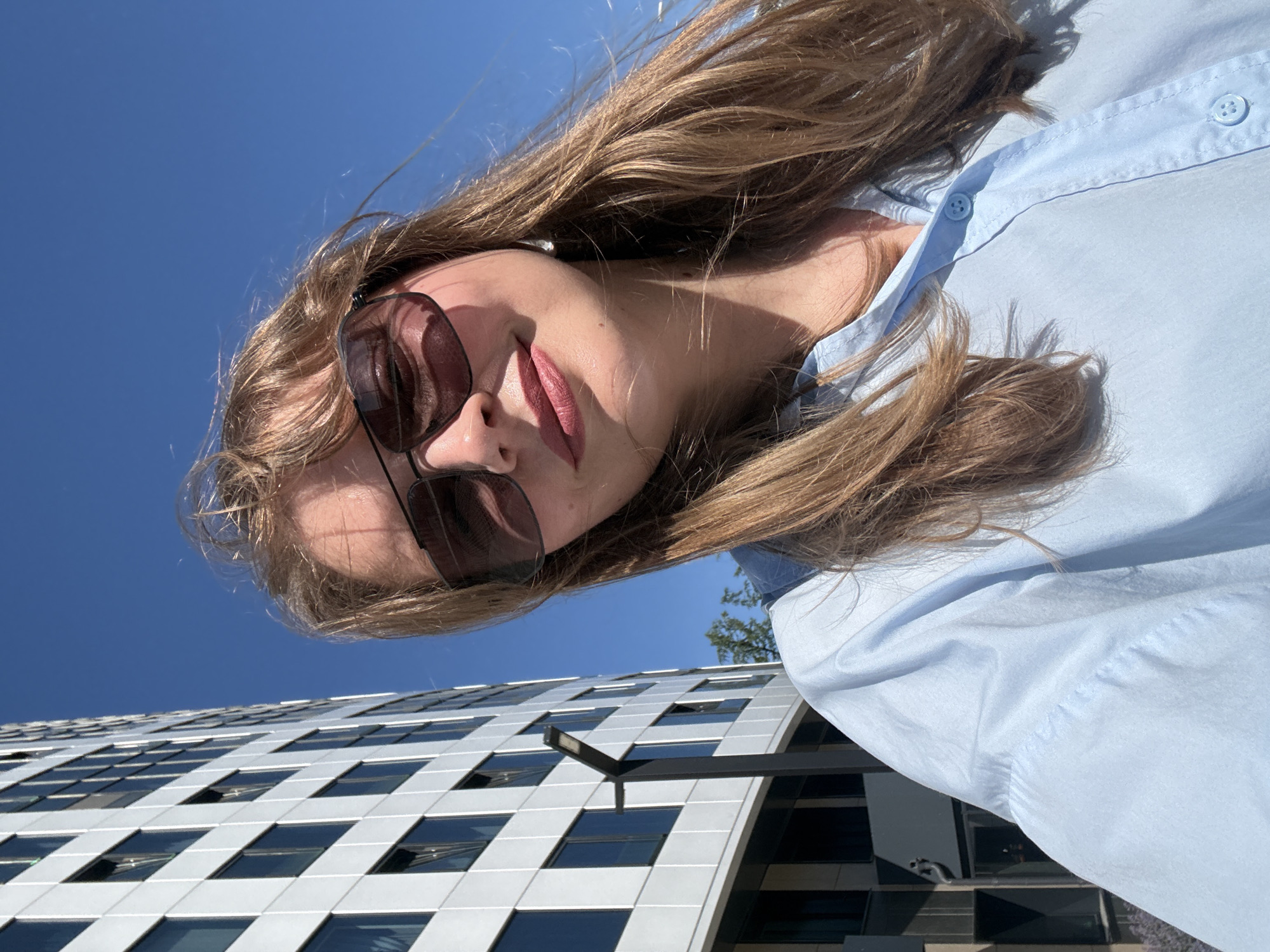 A young Caucasian woman with blonde hair and fair skin takes a selfie outdoors on a sunny day. She is wearing sunglasses and a light blue collared shirt. Behind her, a modern, multi-story building with a facade of white panels and dark windows is visible. A tall, grey lamppost stands in front of the building. The sky is clear and blue. The scene appears to be in an urban environment, potentially in Izhevsk, Russia, given the building's architecture and the clear sky.