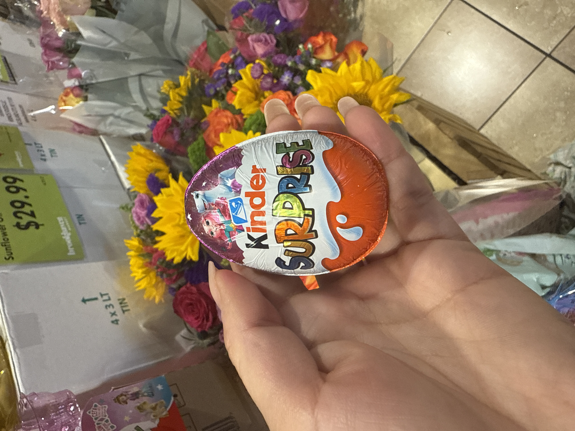 A hand holds a Kinder Surprise egg against a backdrop of a bouquet of flowers and what appears to be a cardboard box with a green price tag indicating "$29.99 Sunflower Oil Brookhaven 4x3LT TIN". The location, based on the "Brookhaven" label, is likely a grocery store in Brookhaven, Darien, United States of America. The image does not show specific people or actions beyond the holding of the Kinder Surprise egg. The environment suggests a purchase or gifting scenario, with the flowers and chocolate treat as possible presents.