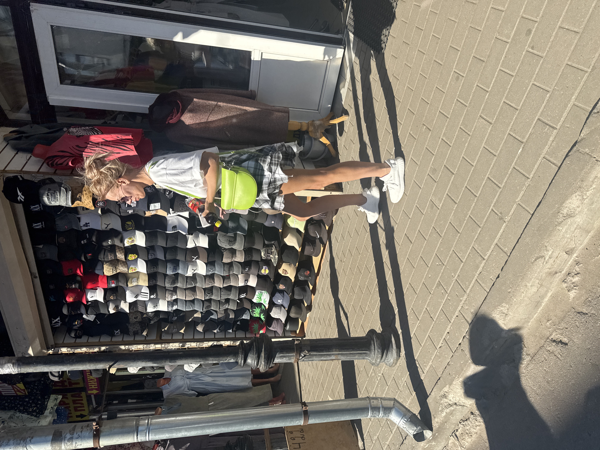 The image shows a young person, appearing to be a teenager, standing on a paved sidewalk in front of a market stall. The person is wearing a white t-shirt, a plaid skirt, and white sneakers. They are carrying a bright green bag over their shoulder and holding a bottle in their hand. The stall is filled with a large display of baseball caps of various colors and designs. Behind the stall, a window is visible, possibly part of a shop or building. The scene is outdoors, with sunlight casting shadows on the ground, suggesting it is daytime. The paving stones indicate an urban or semi-urban environment. The presence of the market stall with merchandise for sale implies a commercial setting. There are no explicit emotional cues visible, and the overall atmosphere appears casual and everyday. Some text is visible on some of the caps and possibly on signs behind the stall, but it is largely illegible due to image resolution and angle. The location context is provided as Saint Petersburg, Russia.