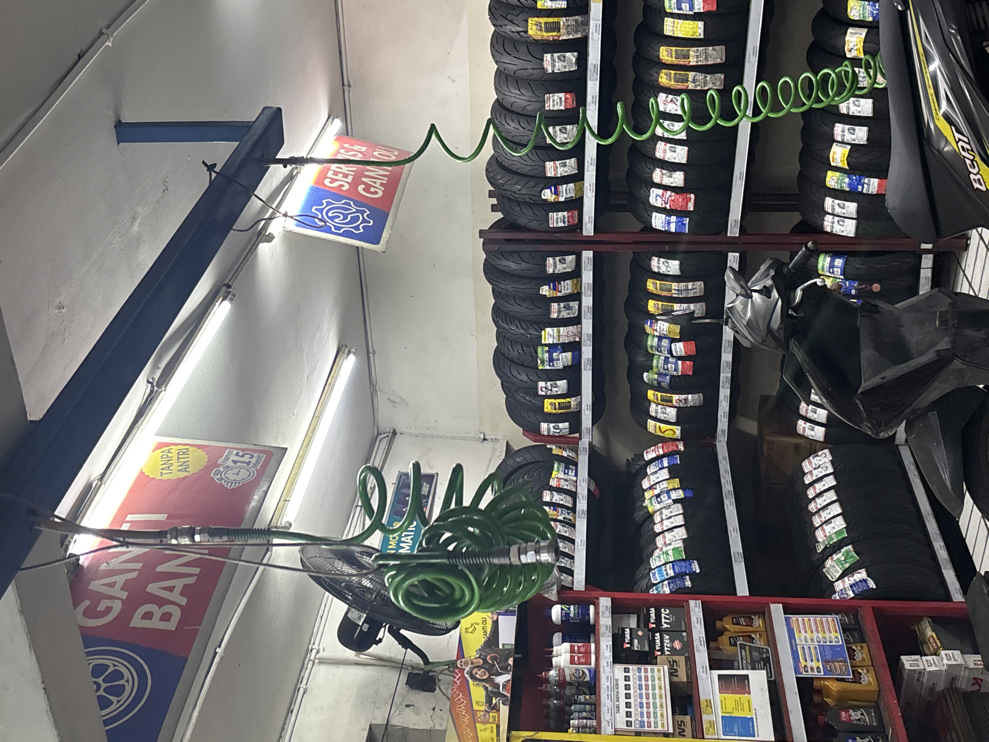 This image depicts the interior of a tire shop in North Jakarta, Indonesia, as evidenced by the Indonesian text on the signage and product labels. The shop appears to be well-stocked with various tires displayed on shelves. In the foreground, a black motorcycle is partially visible, suggesting it might be a customer's vehicle or part of a display. A green coiled hose hangs from the ceiling, likely for air inflation. Various products, such as oil bottles and maintenance supplies, are organized on shelves. The lighting in the shop is provided by fluorescent tubes, illuminating the products and creating a somewhat utilitarian atmosphere. The overall scene is typical of a local auto service and parts store.