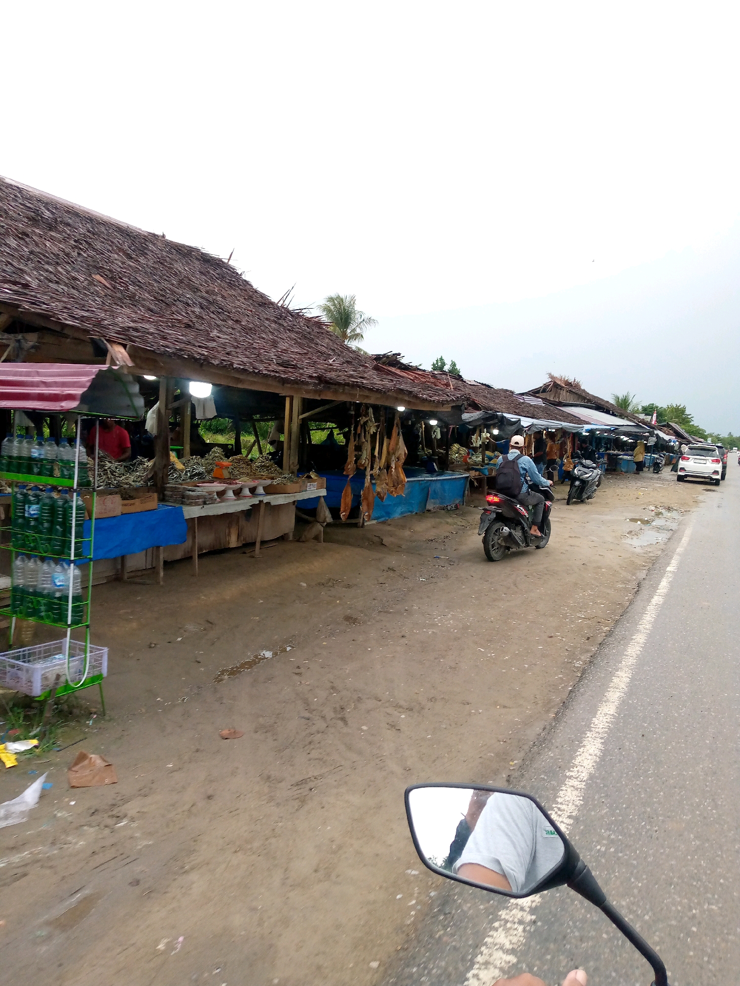 The image depicts a roadside market in Gampongpaya, Indonesia, with several stalls lining the unpaved shoulder of a road. The stalls have thatched roofs supported by wooden posts and appear to be selling dried fish and other dry goods. In the foreground, a person is riding a motorcycle, and the reflection of their arm and shirt is visible in the motorcycle's mirror. On the road, there are other vehicles, including a white SUV and two motorcycles. The environment is overcast, and the ground is a mixture of dirt and gravel, with some puddles of water. The market appears to be a local, informal establishment.