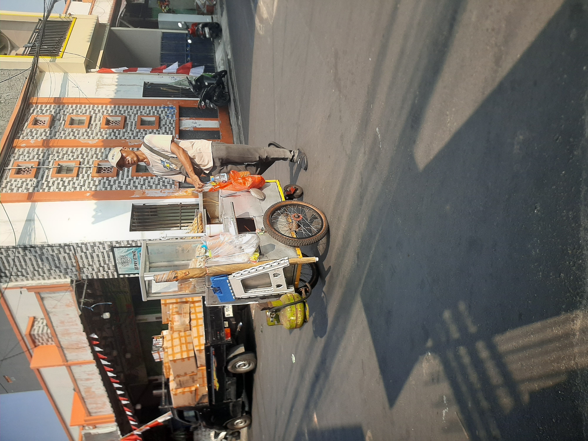 A street vendor walks with his mobile food cart on a sunlit street in Central Jakarta, Indonesia. The vendor, a man wearing a light-colored t-shirt, dark pants, and a baseball cap, is pushing a cart that contains food supplies, a propane tank, and cooking equipment. The cart has a glass display case and a red plastic bag hanging from it. In the background, a black pickup truck is parked with boxes stacked on its bed, and buildings with orange and white facades line the street. Red and white Indonesian flags are visible on one of the buildings. The street is paved and has shadows cast by the buildings and a fence. Two motorcycles are parked on the side of the street. The overall scene depicts a typical urban street in Indonesia during daytime.