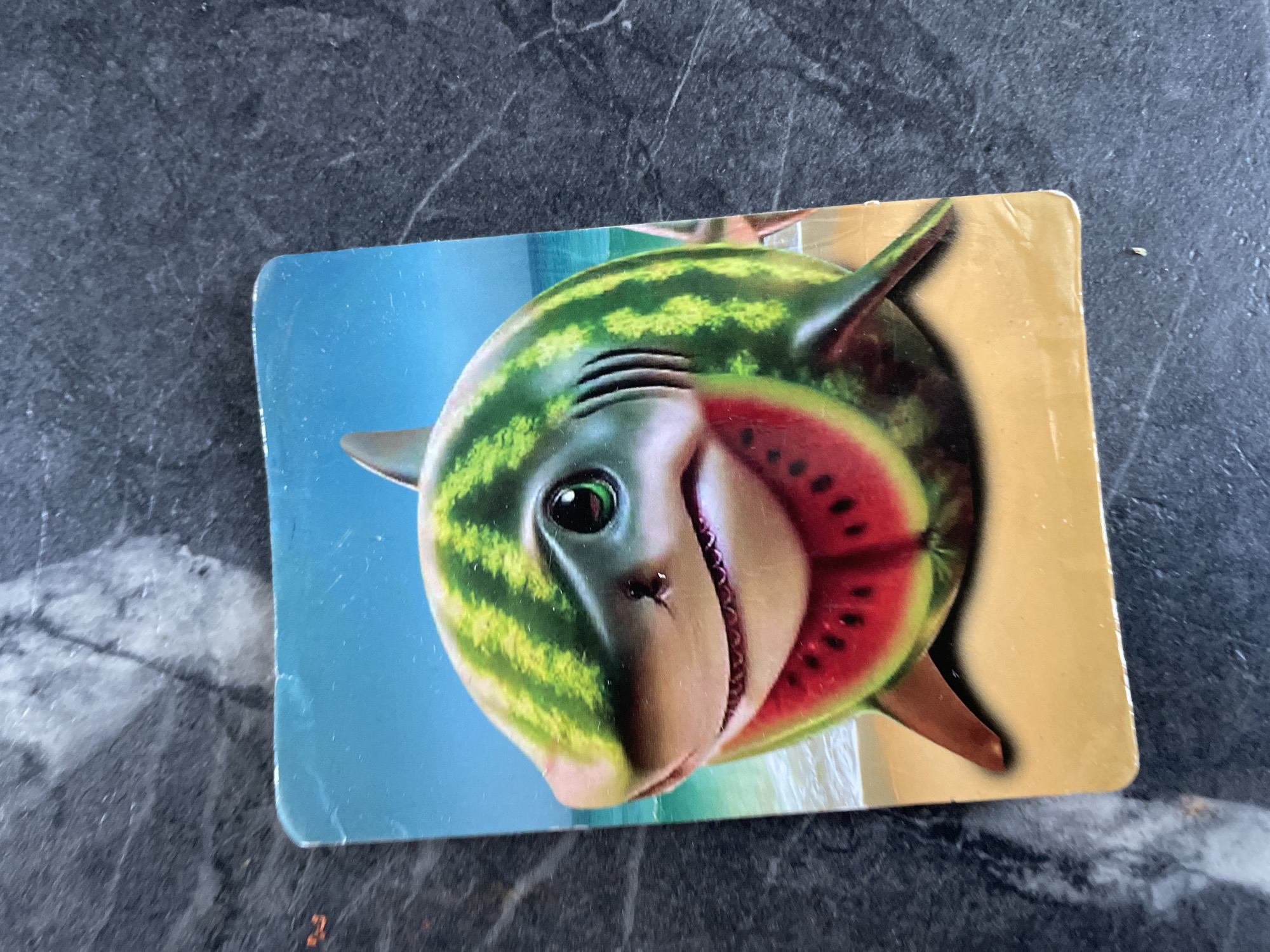 The image, captured in Baranovka, Russia, presents a small, rectangular, laminated card featuring a stylized digital illustration. The central subject on the card is a whimsical creature, appearing as a shark whose body is formed by a watermelon. Its skin displays the characteristic green stripes of a watermelon rind, while its open mouth reveals vibrant red flesh dotted with black seeds. The shark has large, round, green eyes with dark pupils, and faint grey gill slits are visible on its side. It possesses typical dark grey shark fins. The background of the illustration depicts a simplified beach scene, with a clear light blue sky, a calm light blue ocean line with subtle waves breaking on a pale sandy shore. The physical card itself shows minor surface wear and rounded edges. It is positioned on a dark grey, subtly textured surface, likely a countertop, exhibiting faint lighter streaks and a nuanced, granular pattern.