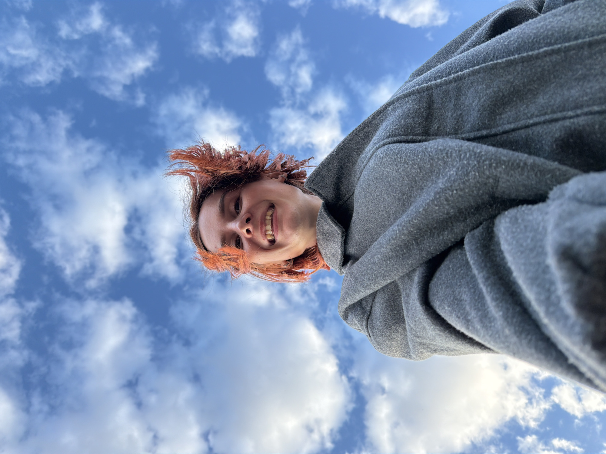 A low-angle shot looking up at a person with red hair, smiling. The person is wearing a gray coat. Behind the person is a blue sky filled with white clouds. The location, Chaikovsky, Russia, cannot be determined from the image alone.