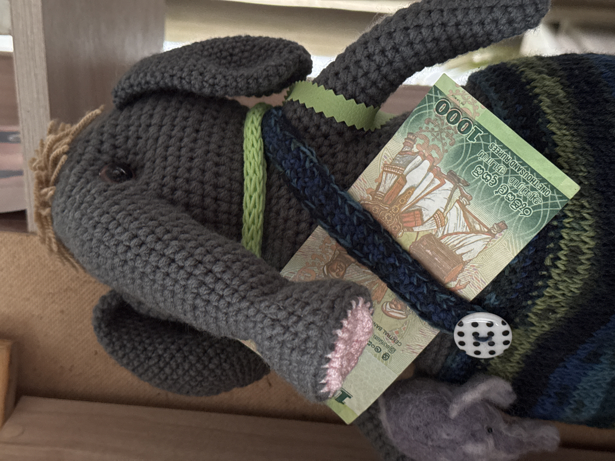 A crocheted gray elephant toy with a One Thousand Sri Lankan Rupee note in its "trunk" area is featured in this close-up image. The elephant is on its side, and a small button with black polka dots is visible to the side of the bill. Parts of the toy are also embellished with green knitted details. The scene does not resemble the city of Ketoshi, Russia, nor any city.