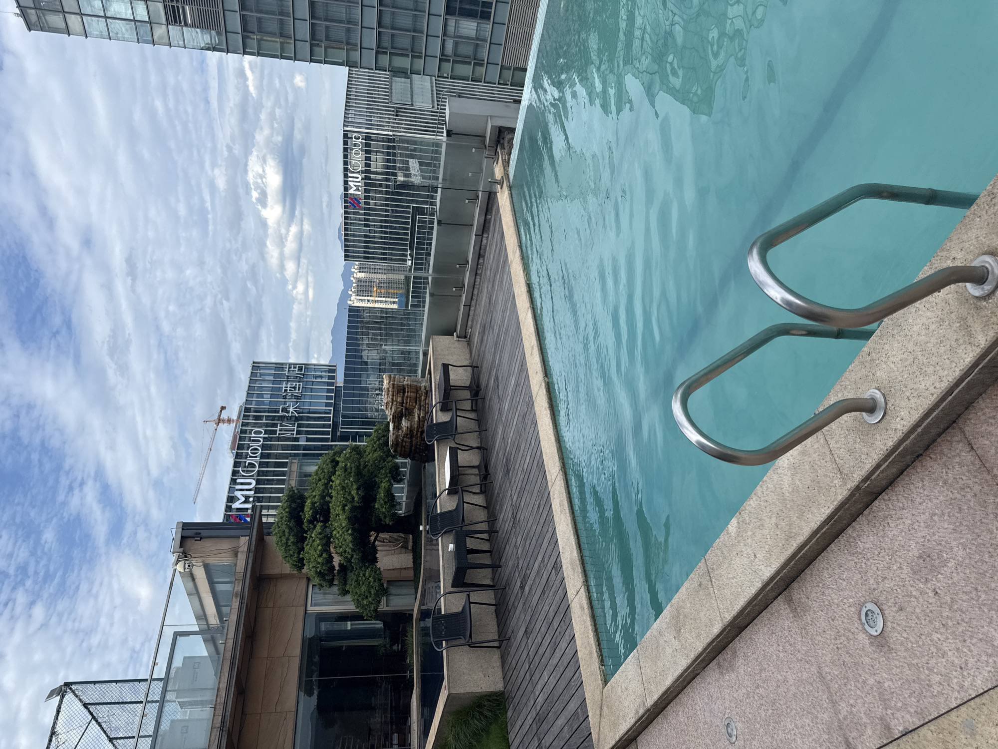 This is a photograph of a rooftop infinity pool in Yiwu, China. The pool is filled with clear turquoise water and has stainless steel handrails. A wooden deck surrounds the pool, furnished with four black chairs and small tables. In the background, modern glass-fronted buildings rise into a cloudy sky. One building displays the "MU Group" logo. The scene depicts a serene urban oasis with a view of the city.