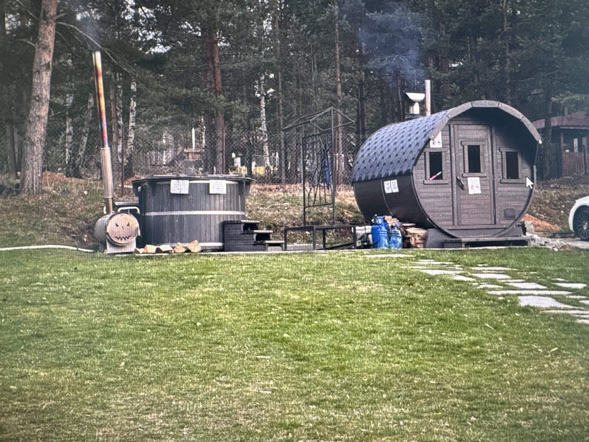 The scene shows a grassy yard with various structures. A wood-fired hot tub with a tall smokestack stands to the left of a barrel-shaped sauna. Between the hot tub and sauna is an open metal structure. To the right of the sauna is a partially visible white vehicle. The background is a dense forest. The location is not explicitly stated.