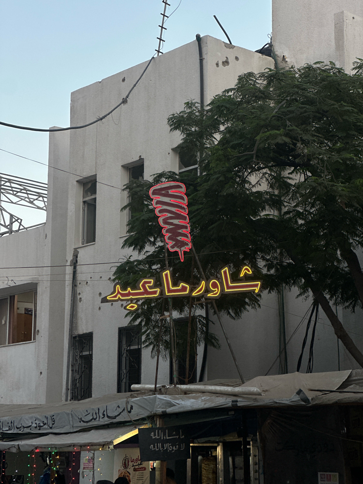 In the city of Gaza, Palestinian Territories, a neon sign featuring a stylized image of a rotating shawarma spit and Arabic script glows brightly against a pale sky. The sign, predominantly red and yellow, hangs in front of a weathered white building. The building's facade shows signs of disrepair, with visible pockmarks and some damaged windows. A large, leafy tree with dark green foliage partially obscures the building on the right side. Below the neon sign, a white awning stretches across the foreground, also showing signs of wear. Underneath the awning, market stalls are visible, with some lights and decorations suggesting commercial activity. The overall impression is of a street scene in a potentially war-affected area, with signs of everyday life continuing amidst visible damage.