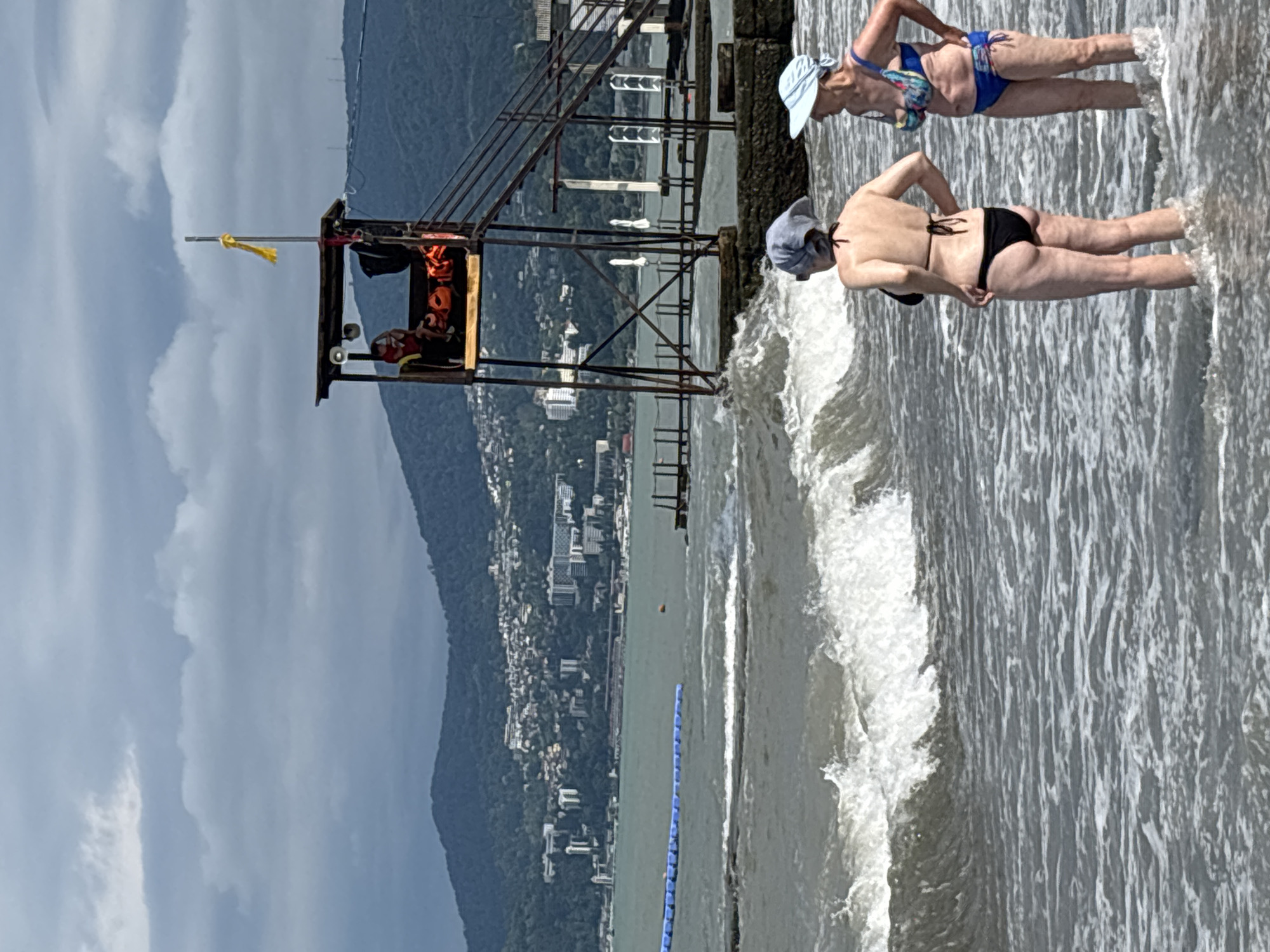 The image depicts a coastal scene in Sochi, Russia, likely on a cloudy day. In the foreground, two women in bikinis stand in the shallow water of the sea, facing away from the camera and towards a distant lifeguard tower. Waves are breaking around their legs. The lifeguard tower, a raised structure with a roof and stairs, stands on a concrete platform extending into the water. A person is visible inside the tower, and several orange life vests are stacked nearby. In the background, the Black Sea stretches towards a coastline dotted with buildings and backed by green, mountainous terrain, characteristic of Sochi's landscape. A blue floating barrier is visible in the water to the left of the frame.