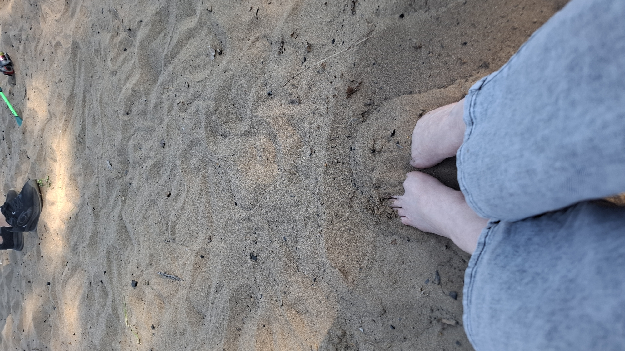 The image shows a sandy surface, likely a beach. A pair of bare feet are visible on the sand, partially obscured by light blue denim jeans rolled up at the cuff. To the left, there is a pair of black shoes, a green fishing rod, and fishing reel. There is no information in the image to confirm the location in the city of Chaikovsky, Russia.