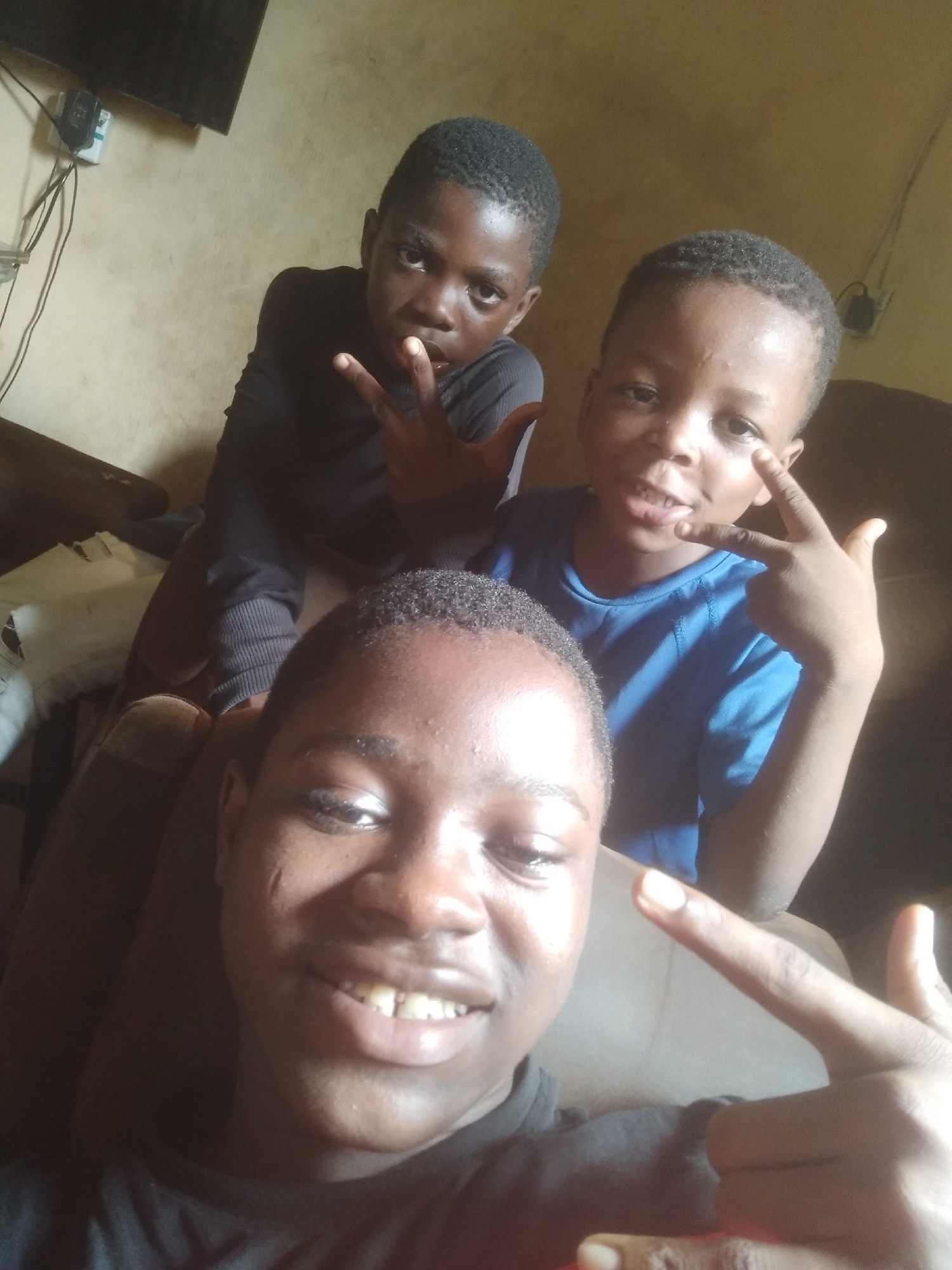 This is a close-up, informal portrait featuring three young males, likely brothers or relatives, posing indoors in Shangisha, Nigeria. The natural lighting suggests the photo was taken during the daytime.
In the immediate foreground, a young man or older boy with dark skin and short, dark hair is smiling widely at the camera, showing his teeth. He wears a black shirt and holds his right hand up, making a peace sign or similar two-fingered gesture.
Behind him and slightly to the left is another young boy, also with dark skin and short dark hair, dressed in a black long-sleeved shirt. He looks intently at the camera with a focused or playful expression, making a "rock on" or similar gesture with both hands.
To the right and slightly behind the foreground subject is the youngest of the three boys, with dark skin and short dark hair. He wears a blue short-sleeved shirt and has a slight smile or smirk. He is making a peace sign or similar gesture with his left hand.
The background features a plain, light-colored wall, possibly cream or beige. In the upper left, a dark television screen, electrical cables, and a white wall-mounted device (likely a power outlet or switch) are visible. Brown furniture, such as a couch or chair, is also present, suggesting a domestic living space. The overall mood is casual and friendly, capturing a cheerful moment among the individuals. No visible text is present in the image.