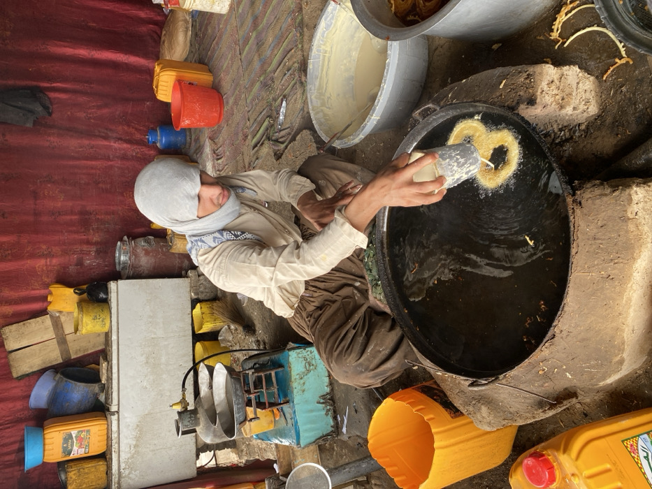 Inside a makeshift food preparation area in Kandahar, Afghanistan, a male individual is actively cooking, likely preparing a sweet pastry. The male, wearing a light-colored long-sleeved shirt, dark loose-fitting trousers, and a light grey head covering with a patterned scarf, is squatting on the ground. He is focused on a large, black, circular pan filled with hot oil, which rests on a crude, mud-built stove structure. With his right hand, he holds a white cylindrical dispenser, pouring light-colored batter into the sizzling oil, forming distinct spiral-shaped pastries that are currently frying.
To his right, a large grey plastic basin contains a quantity of the same light-colored batter. Further right, a metal pot holds already-fried, golden-brown spiral pastries. To his immediate left, a yellow plastic container is on the ground, adjacent to another, similar but uncapped yellow container. In the background on the left, a propane gas cylinder is connected to a small cooking range, topped with a metal pan. Behind this setup, a white, horizontal chest-style freezer/cooler unit is visible, with various plastic containers and buckets (yellow, blue, red) stacked upon and around it. The backdrop consists of a reddish-brown fabric sheet serving as a wall, against which wooden planks lean. Other assorted items, including another blue gas cylinder, additional plastic buckets, and a broom, are visible on the uneven ground, which appears to be a mix of dirt and rudimentary matting. The scene depicts a functional, utilitarian outdoor or semi-indoor cooking operation.