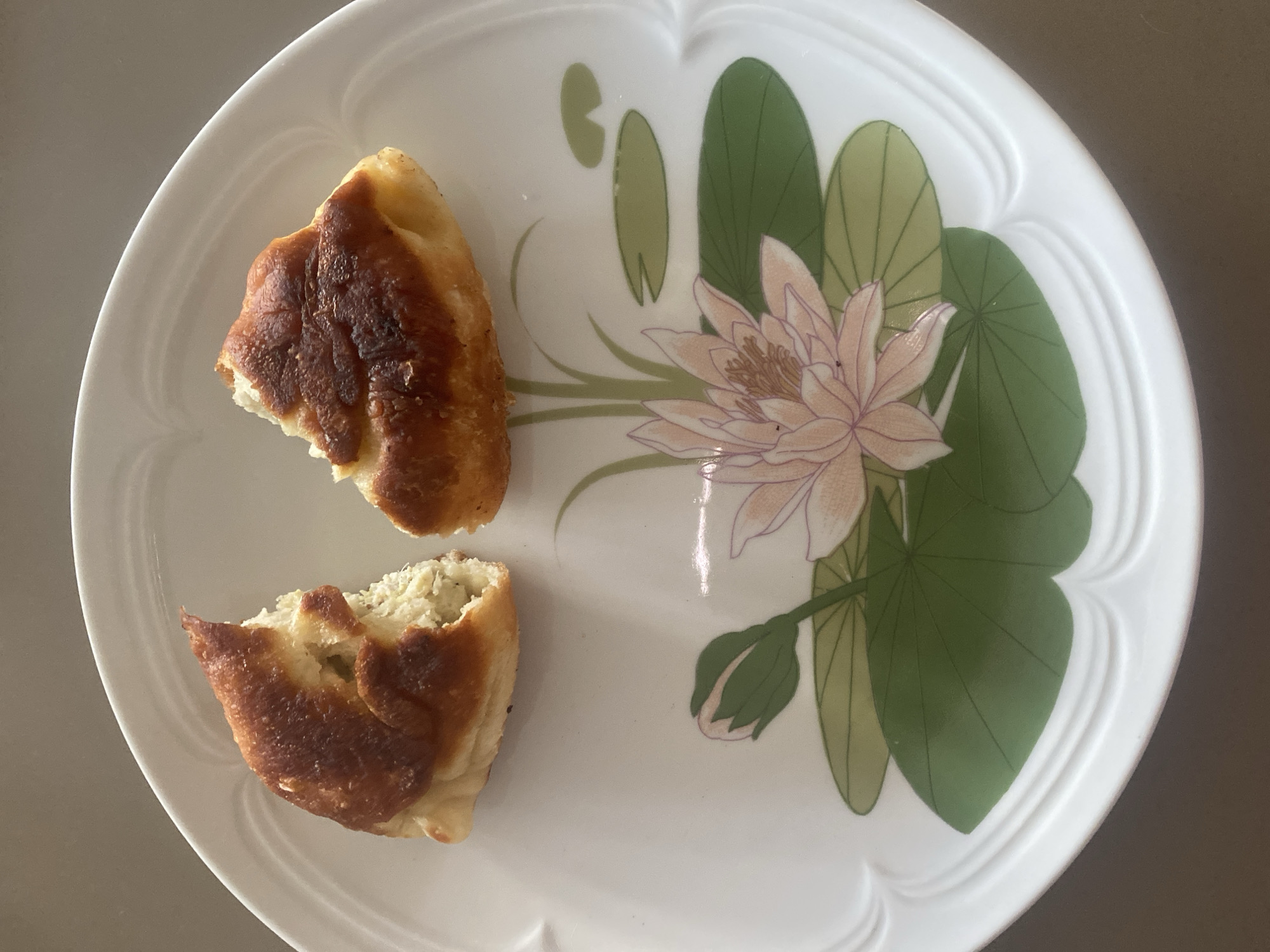 Two pieces of a golden-brown, baked pastry are presented on a white plate. The pastry is broken in half. The plate is decorated with a stylized lotus flower and lily pads in green and pink hues. The background is a neutral, mottled surface. The location is not specified in the image.