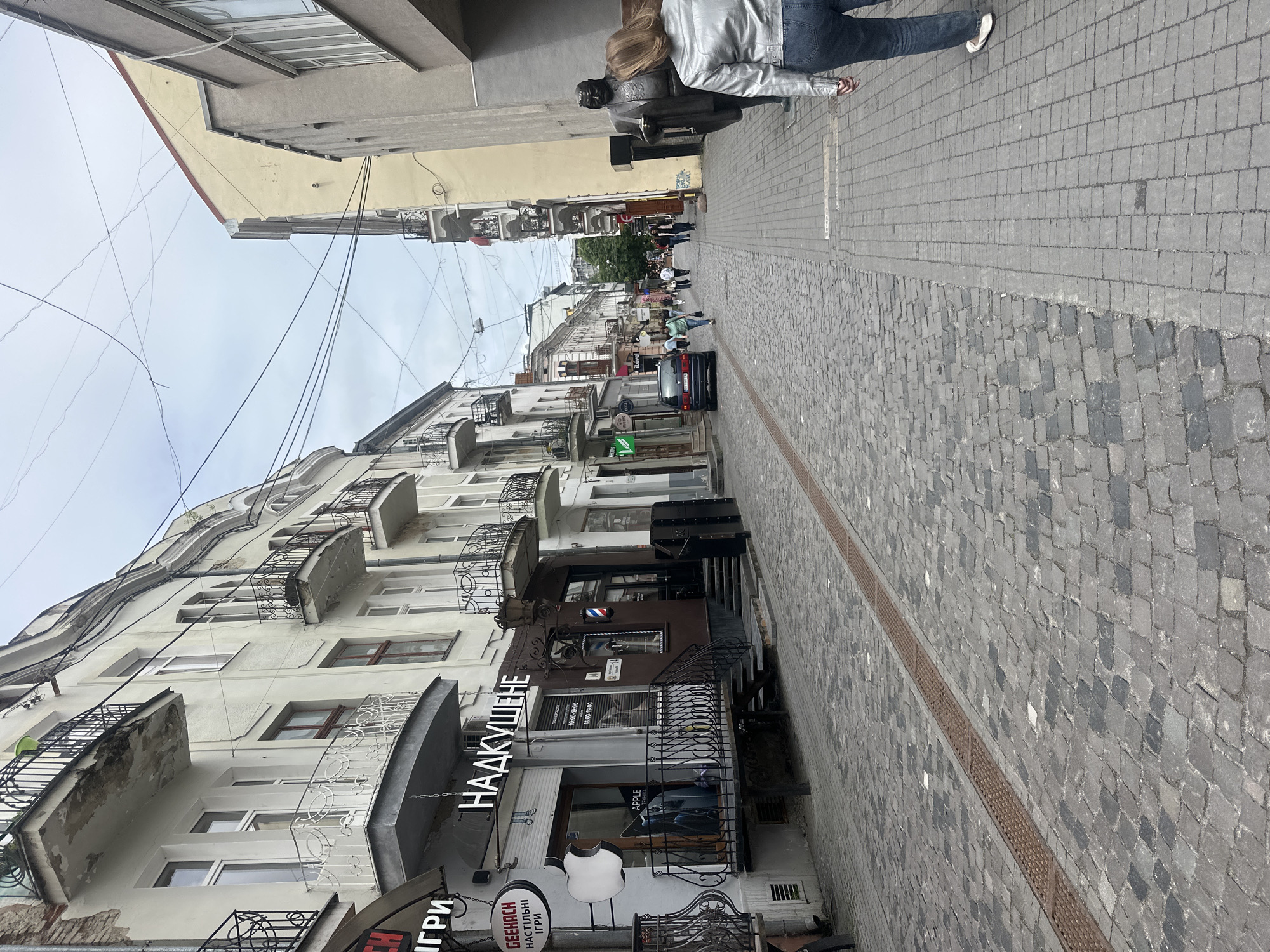 This is a street scene in Ternopil, Ukraine. The street is paved with cobblestones and lined with buildings on both sides. In the foreground, a woman in a silver jacket and jeans is walking away from the camera. To her left, there is a statue of a man in a suit. The street continues into the distance, where other people can be seen walking. There are shops and businesses on the ground floor of the buildings, including a barber shop with a barber pole. Overhead, there are numerous electrical wires strung across the sky. The buildings are multi-story and appear to be a mix of old and newer architecture. The sky is overcast.