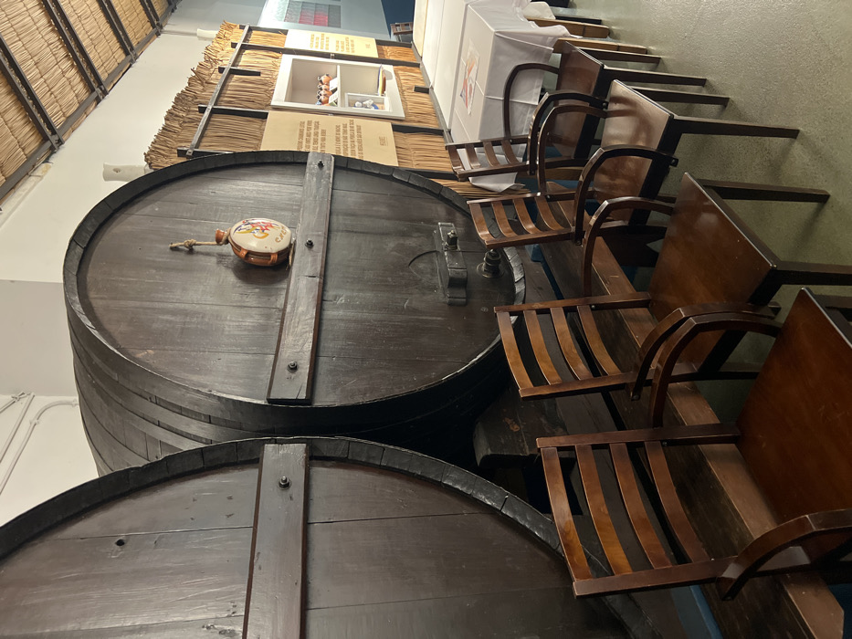 An interior view depicts two large, dark-stained wooden barrels on wooden stands, occupying the left and central portions of the scene in Coruche, Portugal. The barrel on the right features a brass spigot near its base and a multicolored ceramic flask suspended by a cord on its face. In front of these barrels, a row of six empty dark brown wooden chairs with slatted backs and armrests is arranged. To the right, multiple tables covered with white tablecloths are visible. The background wall is paneled with thatched or reed material, displaying informational posters with Portuguese text and a white alcove containing small, colorful bird figurines and other items. The ceiling is also constructed from similar thatched or reed material. No individuals are present.