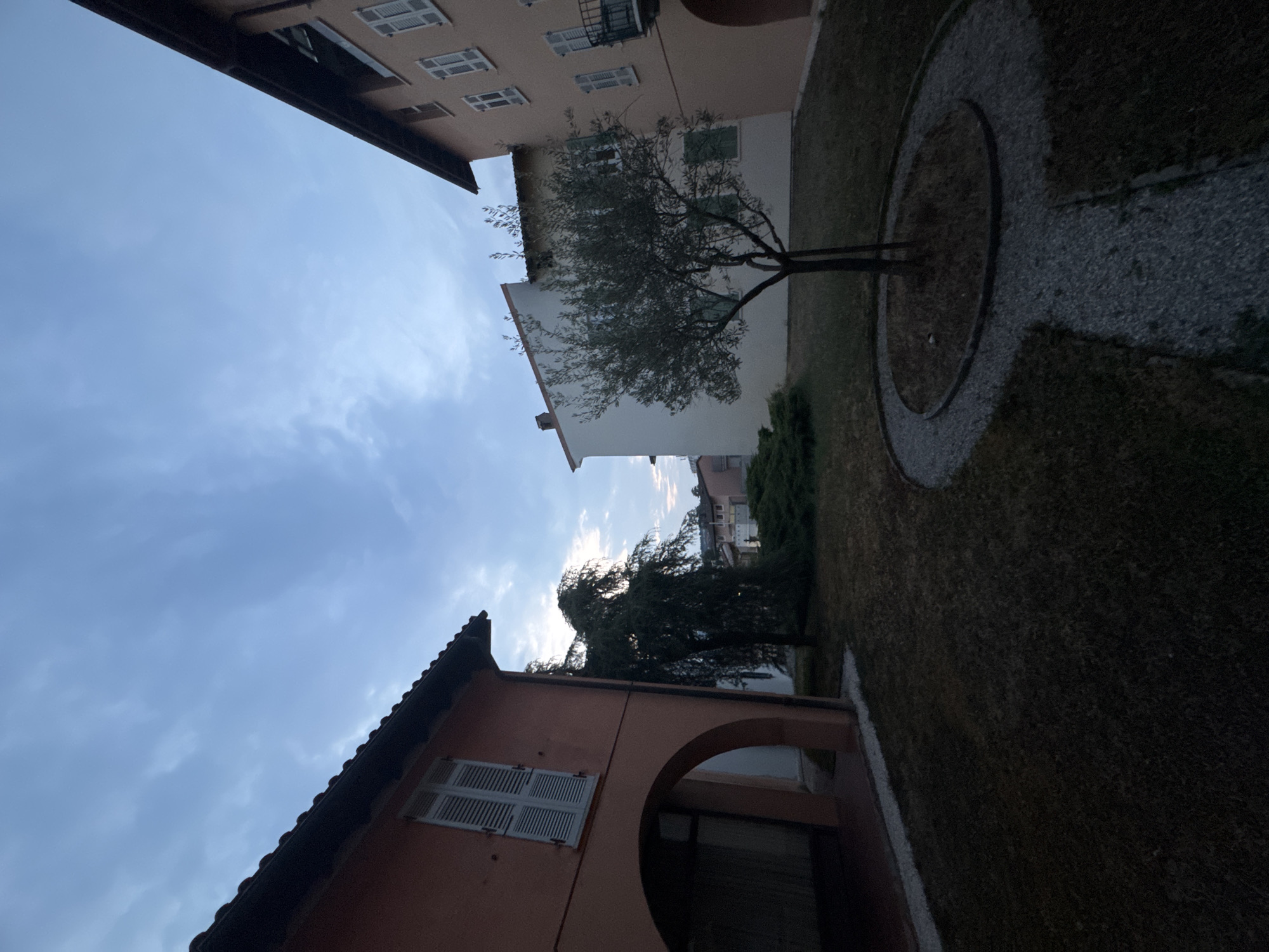 The image shows a view of a courtyard or garden area with buildings on either side and in the background. The sky is overcast with some lighter clouds. In the foreground, there is a lawn with a gravel pathway leading to a small tree. To the left is a building with a salmon-colored facade, featuring a large arched opening on the ground floor and a window with white shutters on the upper floor. To the right is a light-colored building with multiple windows and shutters, and a balcony. Further in the background, more buildings are visible. The scene appears to be set in the late afternoon or early evening, given the lighting conditions. The location is identified as Balerna, Switzerland.