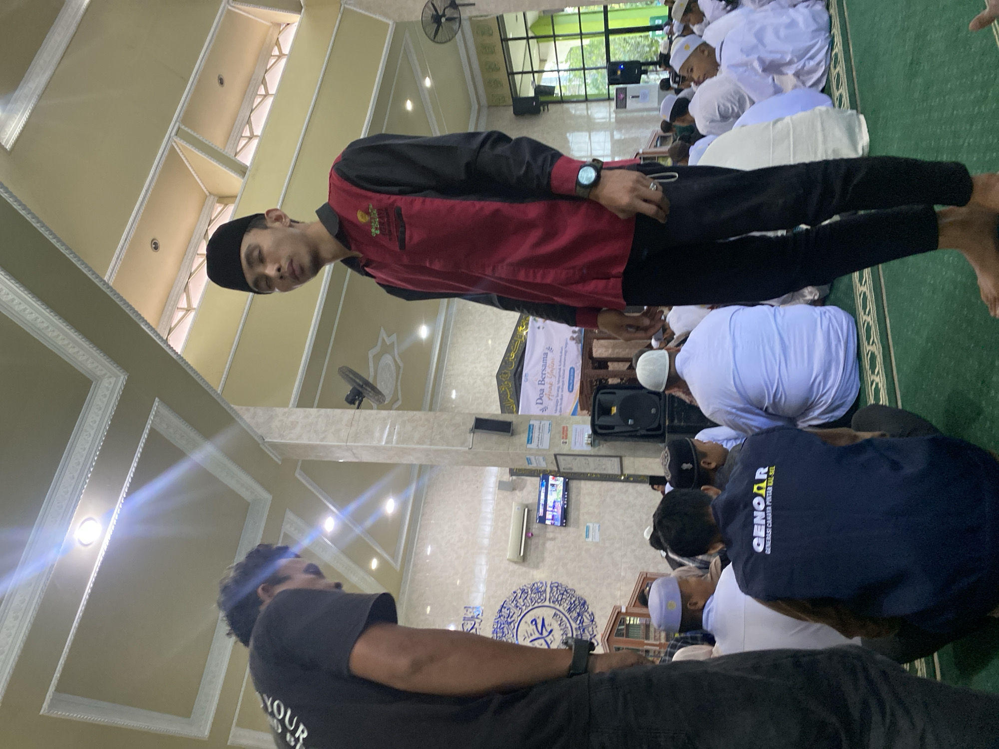 In Banjarbaru, Indonesia, a group of young men and boys are gathered on the green carpeted floor of what appears to be a mosque. The room has high ceilings with ornate white decorative molding and recessed lighting. The walls are tiled with a beige, marbled pattern.
In the foreground, a young man wearing a black peci (a cap common in Indonesia) and a red and black collared shirt stands facing the camera. He has a watch on his left wrist. To his left, another young man in a black t-shirt is partially visible, with text on his back that reads "YOUR".
The other individuals in the scene are mostly seated, many of them wearing white peci or skullcaps and white clothing. Some are facing away from the camera, looking towards the front of the room. One individual in the middle ground is wearing a dark blue vest with white text that says "GENOAR GENERASI CERDAS PINTAR KAL-SEL".
There are several objects in the environment, including air conditioning units mounted on the wall, speakers, a television screen, and banners with text in Indonesian. A large blue circular design with Arabic calligraphy is visible on the wall behind some of the seated individuals. The overall atmosphere suggests a gathering or event taking place in a religious or community setting.