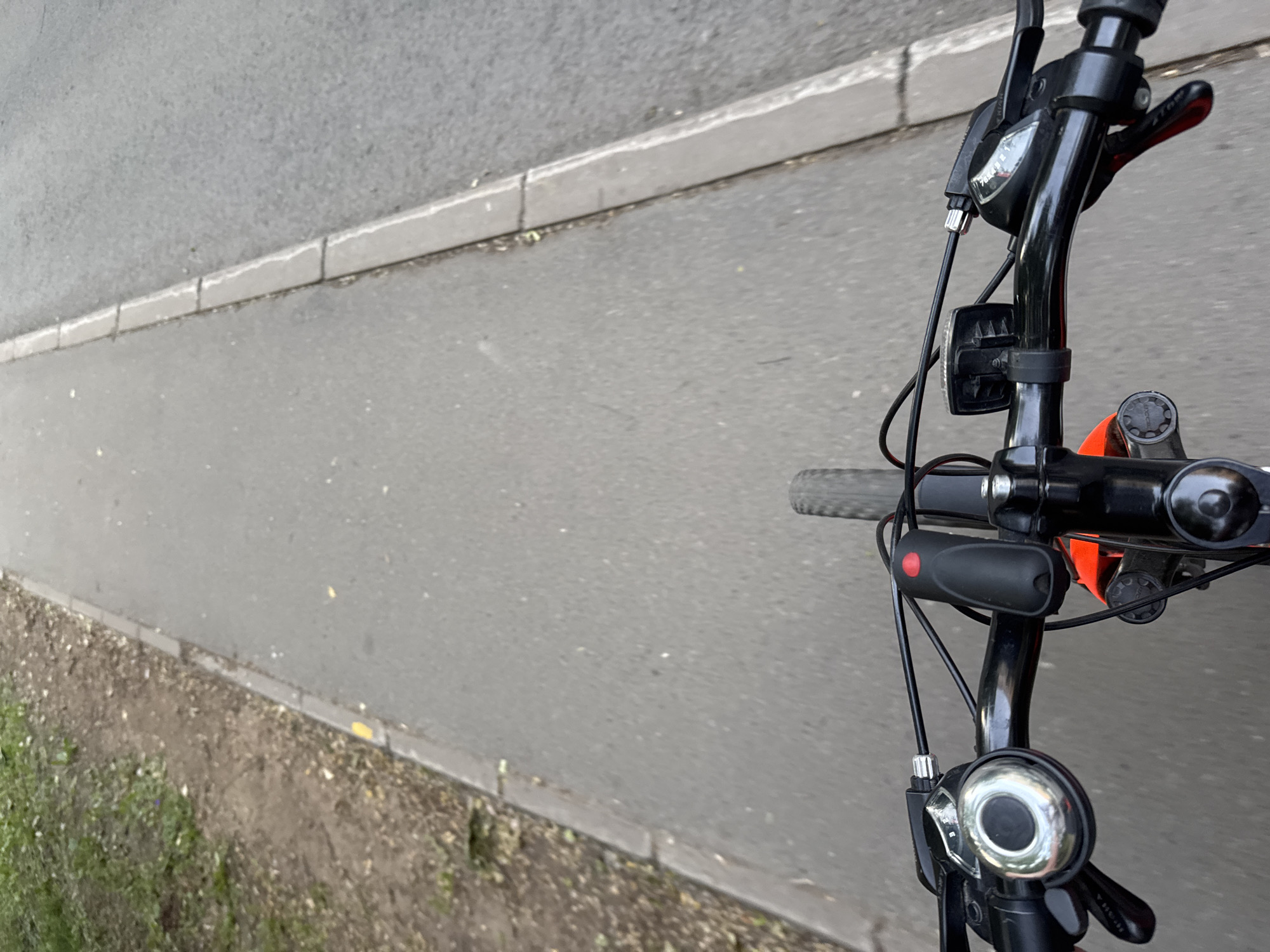 The image shows a first-person perspective of someone riding a bicycle on a paved path next to a grassy area. The handlebars of the bicycle are visible in the foreground, with a bell, gear shifters, and a red light attached. The path appears to be a dedicated bicycle lane or sidewalk, bordered by a concrete curb. The environment suggests an outdoor setting, possibly a park or a tree-lined street. No specific landmarks or identifying features of Saint Petersburg are visible.