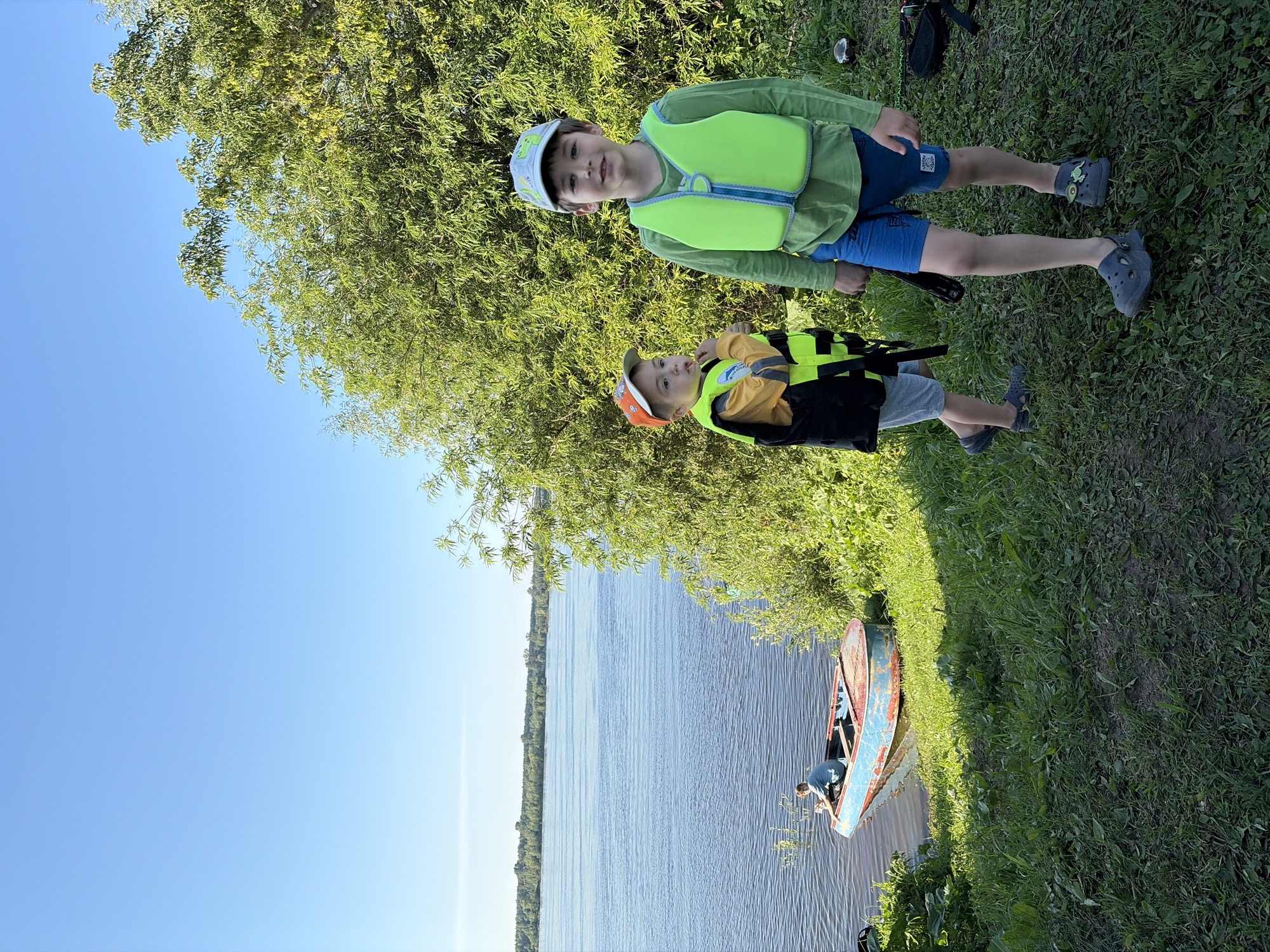 The image captures a sunlit scene outdoors, likely in or near the village of Olkhovkа, Russia, given the presence of a wide river and lush vegetation. In the foreground, two young boys are standing on a grassy embankment overlooking the water. The boy on the right, older of the two, wears a lime green long-sleeved shirt, blue shorts, and a bright green life vest. He also sports a light-colored baseball cap. The younger boy, to the left, is dressed in a yellow long-sleeved shirt, grey shorts, a black vest, and an orange baseball cap. He is holding something to his mouth, possibly a snack. Both boys are wearing casual footwear.
To the left of the boys, near the water's edge, is an old, weathered boat painted in red and blue. A person, whose face is not clearly visible, is seated inside the boat, seemingly attending to something. The river is wide and calm, reflecting the clear blue sky. Beyond the river, a distant tree line marks the horizon. The vegetation on the embankment is dense and green, with a large tree providing shade in the background. The overall atmosphere is peaceful and suggests a recreational outing by the water.