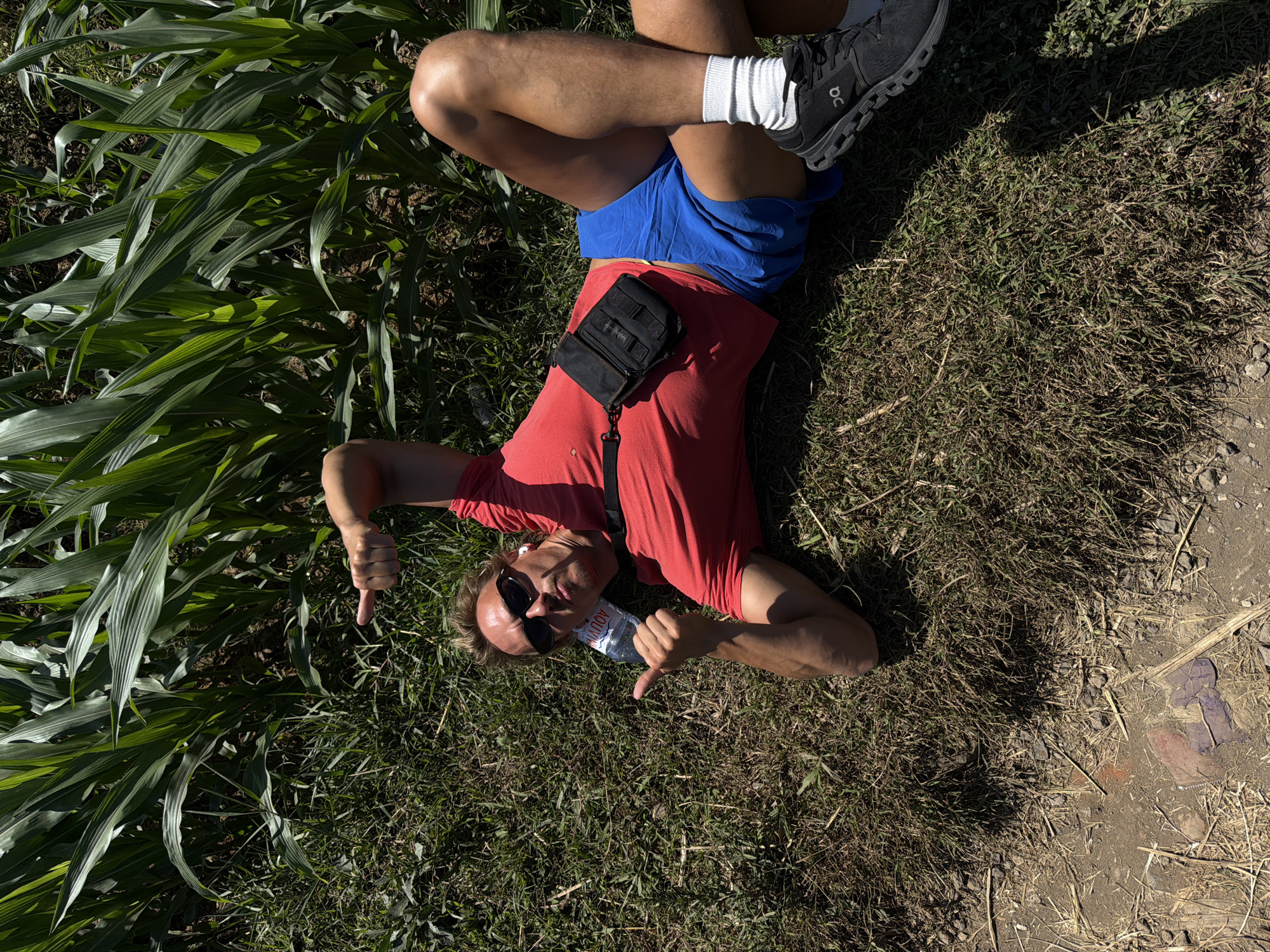 A man with blonde hair and sunglasses is lying on his back in the grass next to a cornfield. He is wearing a red t-shirt, blue shorts, white socks, and black athletic shoes. A small black pouch is attached to his t-shirt. He is making hand gestures with both hands, pointing with his left index finger and giving a thumbs-up with his right hand. A bottle of water is on the ground beside him. The scene is outdoors on a sunny day, with tall green corn stalks visible in the background. The location is within the city of Dampit, Indonesia.