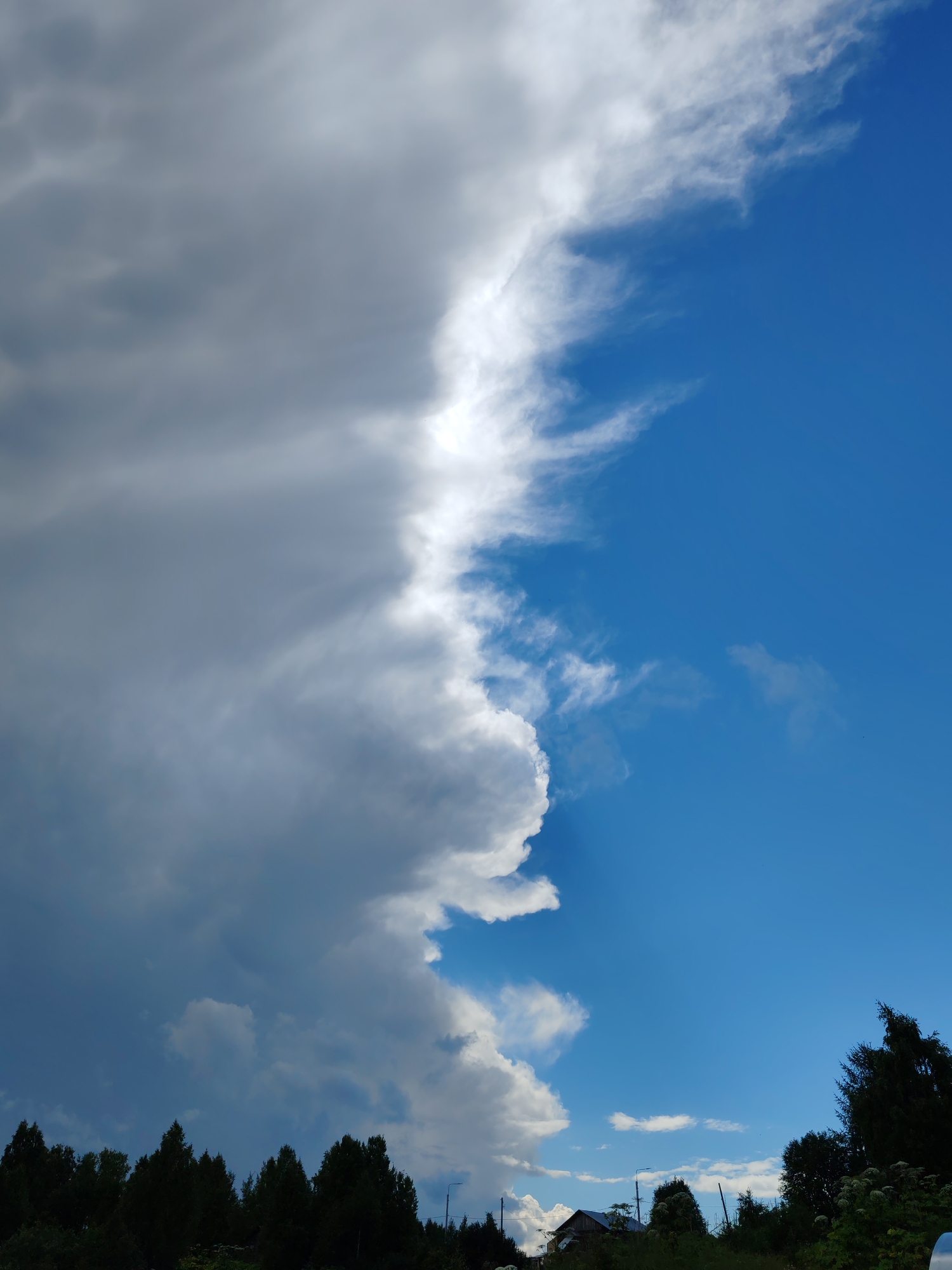 The image shows a dramatic sky with a stark division between dark, heavy storm clouds on the left and bright blue sky with wispy white clouds on the right. This meteorological boundary runs diagonally from the top left to the bottom right of the frame. Below the sky, a dense line of dark green coniferous trees forms a silhouette against the clouds and sky. In the lower portion of the image, a small, rustic house with a dark roof is visible nestled amongst the trees, and a few scattered smaller structures and utility poles are also present. The overall impression is of an impending storm or the aftermath of one, set against a rural landscape in Чухлэм, Russia.