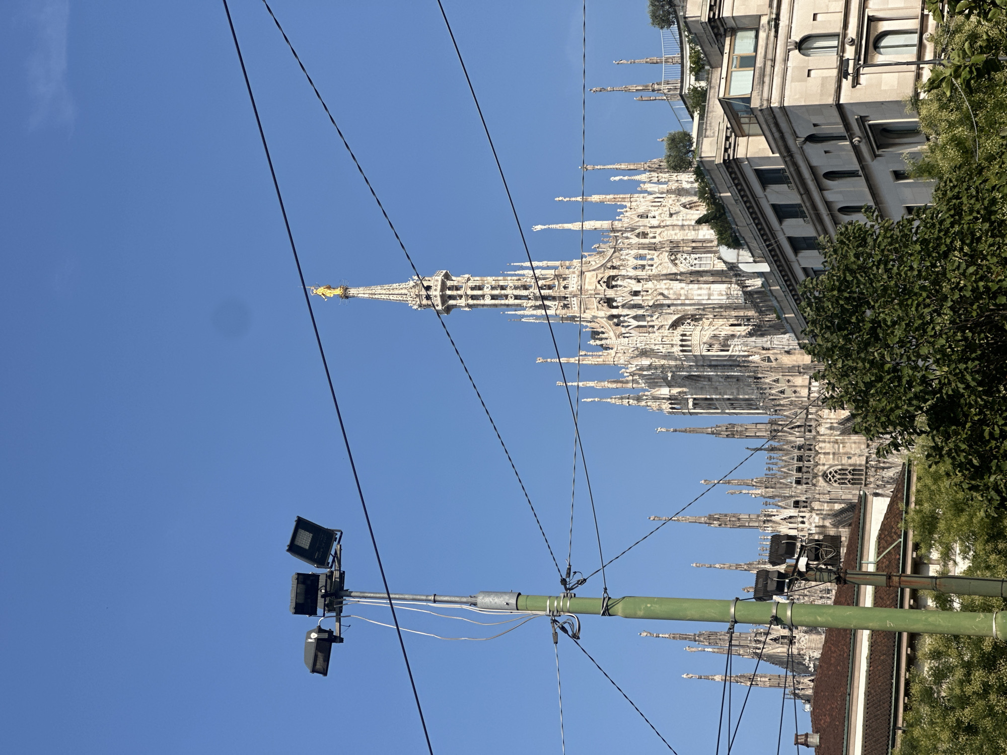 The image shows a view of the Duomo di Milano, a cathedral located in Milan, Italy. In the foreground, a green lamppost with three lights is visible, along with overhead power lines. Behind the lamppost and power lines, the intricate Gothic architecture of the Duomo is prominent, with its spires and detailed facade. A golden statue of the Virgin Mary, known as the Madonnina, is visible atop the highest spire. The sky is clear and blue. Several buildings, some with trees and greenery, are situated between the lamppost and the Duomo. The image appears to be taken from street level, looking up towards the cathedral.