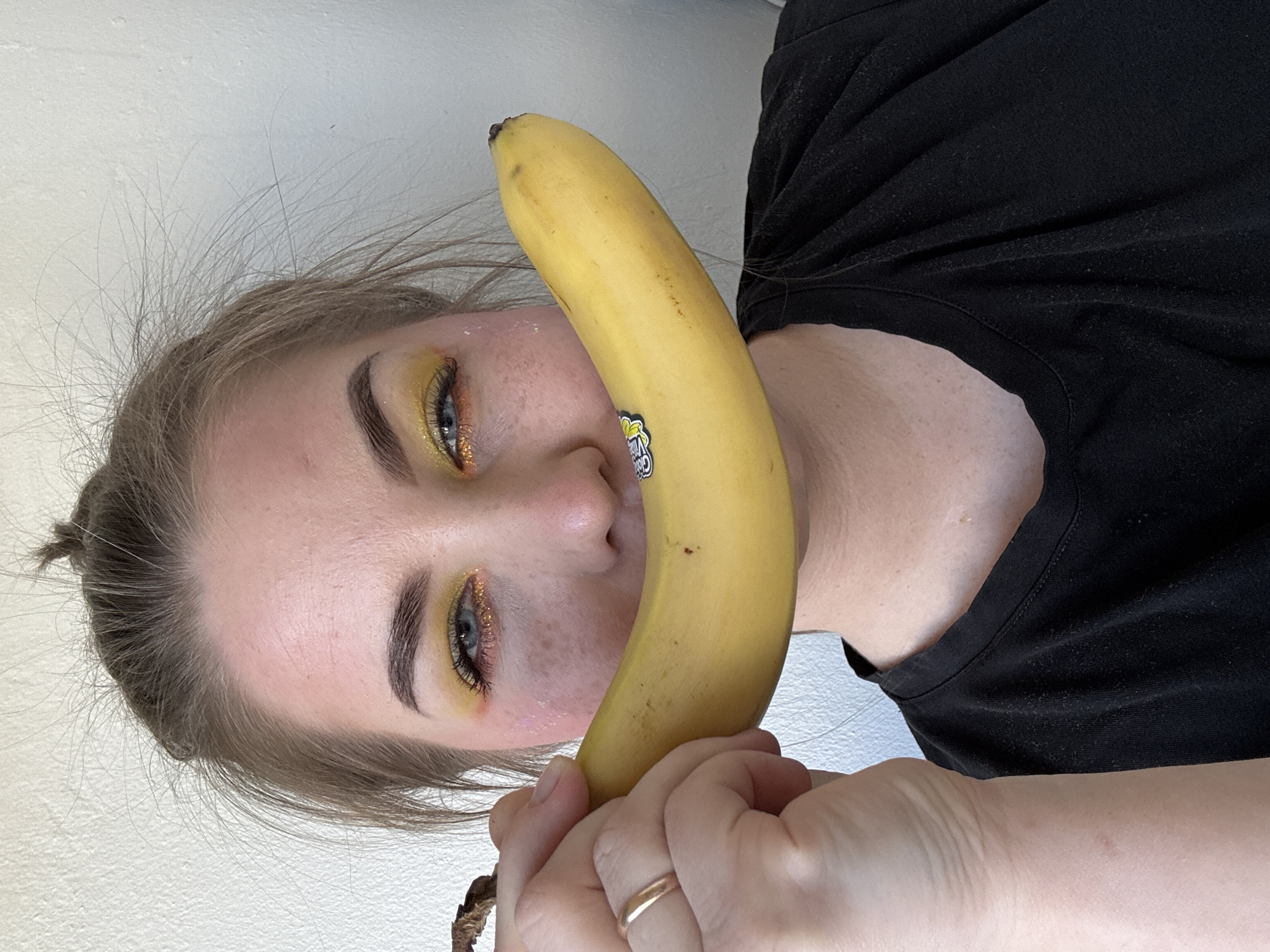 A fair-skinned person with blonde hair partially tied back holds a yellow banana against their face. The banana is positioned vertically, with the top end near the person's forehead and the bottom end held in their hand. A Chiquita sticker is visible on the banana near the person's nose. The person has yellow and orange eyeshadow and is wearing a black shirt. The background is a plain white wall. There is no evidence to suggest the image was taken in the city of Chaikovsky, Russia.