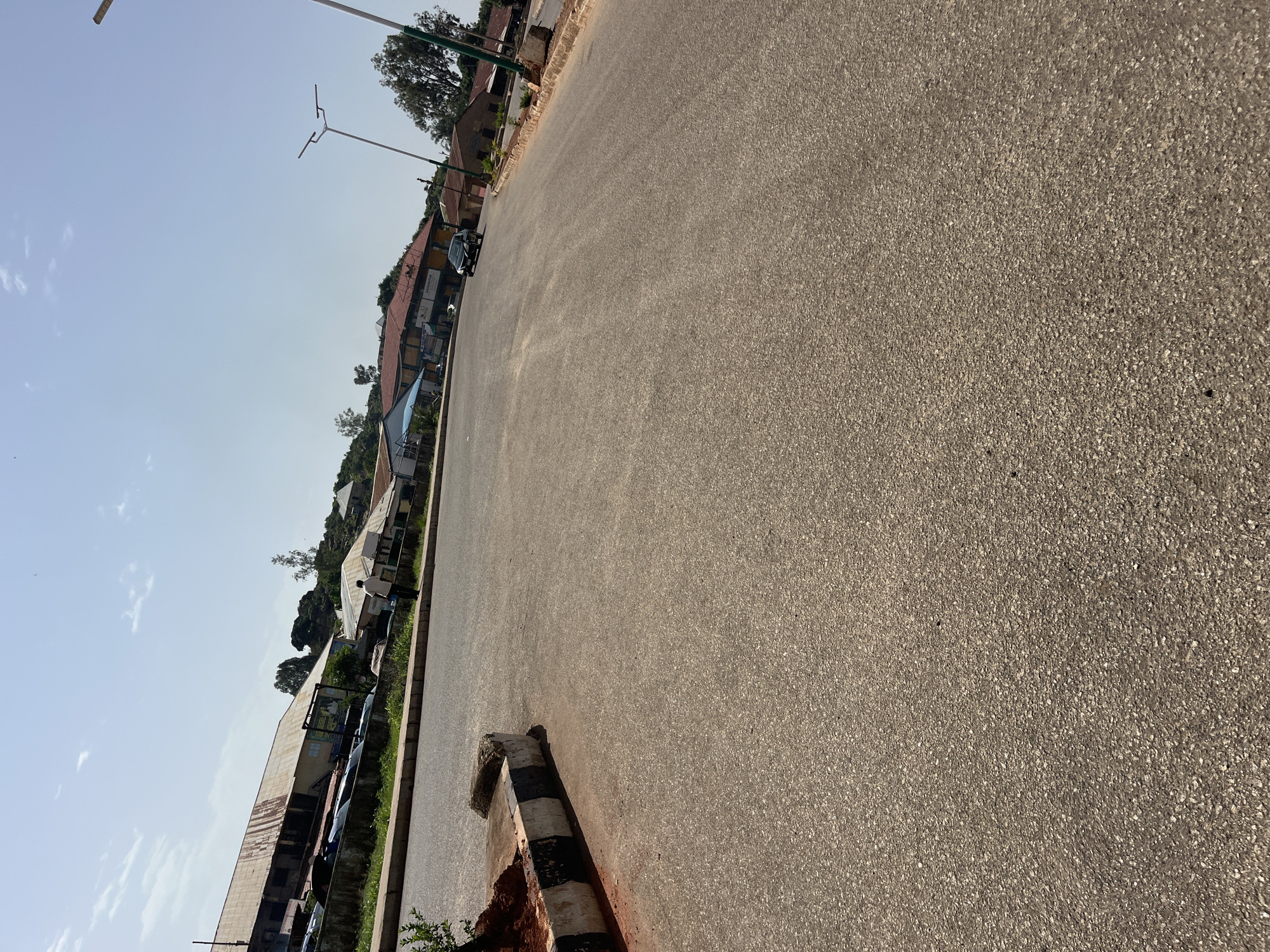 In Bukuru, Nigeria, a wide asphalt road occupies the foreground, stretching diagonally from the bottom left to the upper right of the frame. The road surface is textured with small, light-colored stones embedded in darker asphalt. Along the left side of the road, a raised curb is visible, painted with alternating black and white stripes. Beyond the curb, a patch of dry, reddish-brown earth is visible, with a few small green plants.
To the left of the road, a row of buildings lines the street. The first building appears to be a long, low structure with a rusty corrugated metal roof and a light-colored facade, possibly with windows. Further down the street, other buildings with varied rooflines and colors are visible, suggesting a commercial or residential area. Some of these buildings have signs, and several parked vehicles are present along the roadside. Trees and vegetation are scattered throughout the background, with some rising up a gentle slope behind the buildings.
On the right side of the road, more buildings are visible, including structures with reddish-brown roofs and light-colored walls. Streetlights with modern designs are positioned along the road. A dark-colored car is driving away from the viewer on the road. The sky above is clear and bright blue, with a few wispy white clouds. The overall impression is a street scene in a developing urban or semi-urban area under a bright, sunny sky.