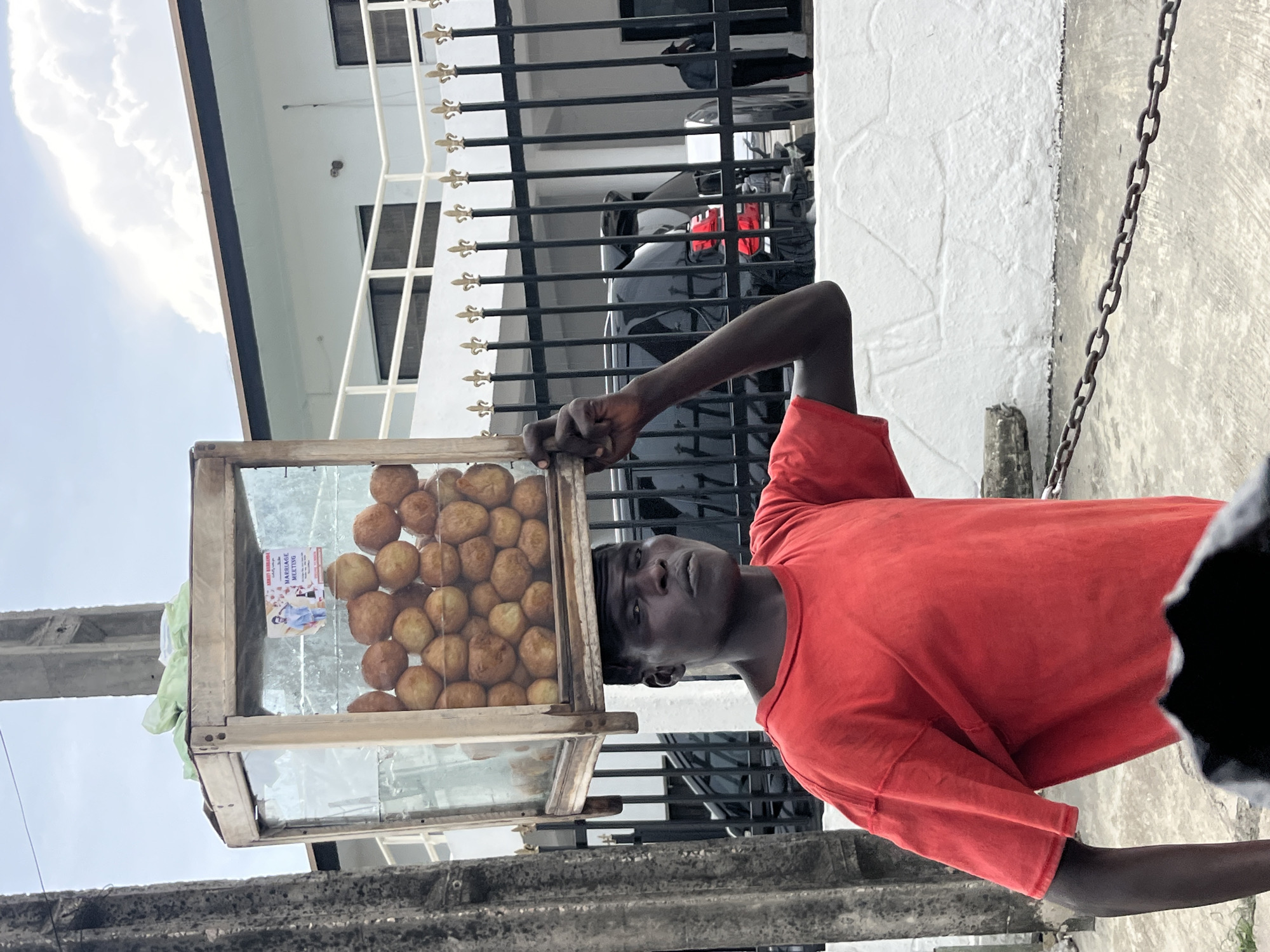 A street vendor in Itirin, Nigeria, balances a glass case filled with fried dough balls on his head. He is a dark-skinned man wearing a red t-shirt. The case contains approximately 40-50 golden-brown, round pastries and a small poster. The vendor is positioned next to a building with black metal bars over windows and a gray car parked behind the bars. A gray cement sidewalk and chain fence are visible on the right. A concrete post is seen on the left. The sky is partly cloudy.