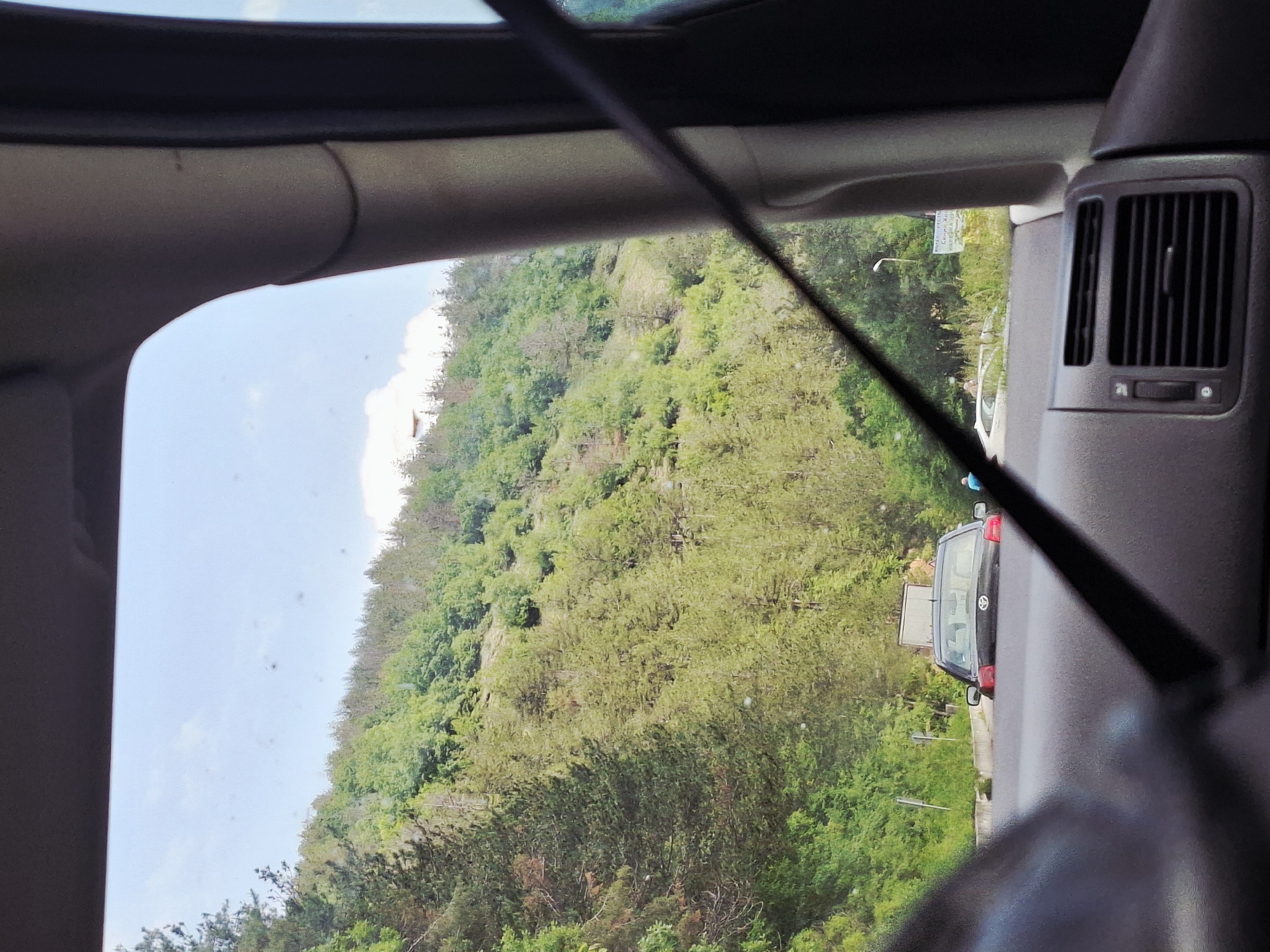 This image captures a view from inside a vehicle, looking out a side window at a landscape. The perspective is rotated 90 degrees clockwise, suggesting the camera was held vertically.
In the immediate foreground, parts of the car's interior are visible: the dark upper trim along the window frame, a section of the car's pillar, and a diagonal dark strap, likely a seatbelt, cutting across the frame from the top left towards the bottom right. On the far right, a car air vent is clearly visible, set into the interior paneling.
Outside the window, a bright, clear sky with hints of white clouds fills the upper portion of the frame. Below the sky, a densely forested, lush green hillside or mountain dominates the left side of the view, appearing steep and verdant.
On the right side of the exterior view, a road is visible. A dark-colored hatchback or small SUV is parked or moving slowly along this road, facing away from the viewer. A faint logo, possibly Toyota, is discernible on its tailgate. Near the roadside, there's a white rectangular sign with partially visible, illegible text (likely Cyrillic, given the location). A street lamp pole also stands by the road. A small blue figure, possibly a person, can be seen in the distance near the road and the parked car.
The scene depicts bright daylight conditions, suggesting a sunny day. There are no clear indicators of specific activities other than travel by car and observation of the scenery. The setting, with its mountainous terrain and dense greenery, is consistent with the location of Hvoina, Bulgaria.