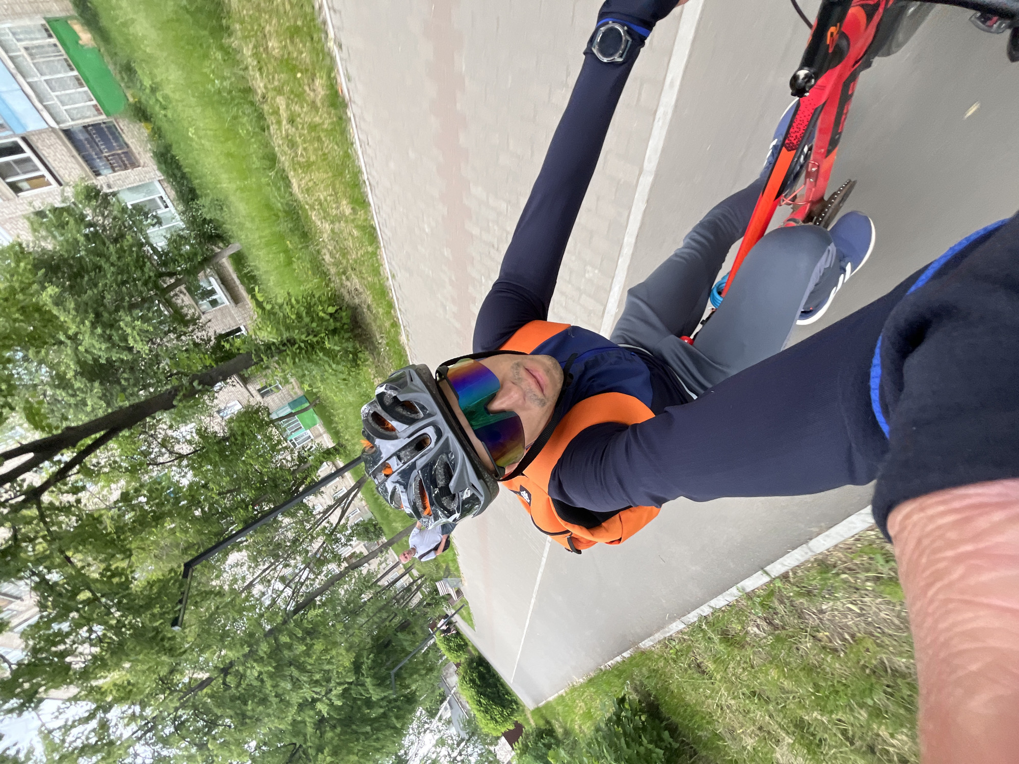 A man is riding a bicycle on a paved path in Chaikovsky, Russia. He is wearing a helmet, sunglasses, and athletic clothing, including an orange backpack. The path appears to be a dedicated bike lane. In the background, there are trees, grass, and apartment buildings, indicative of a residential area. Another person is visible further down the path. The lighting suggests it is daytime, possibly overcast.
