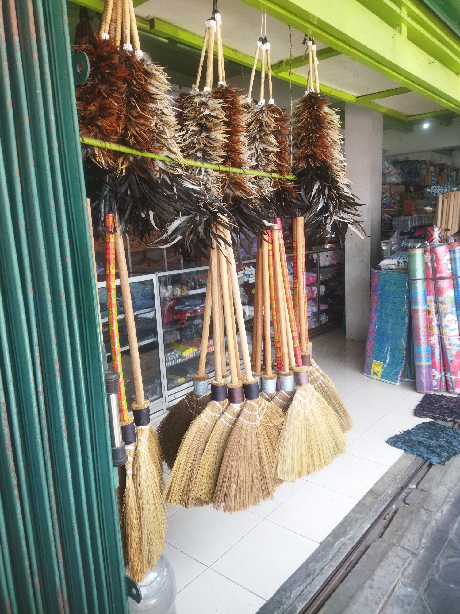 In the city of Wonogiri, Indonesia, an outdoor market stall displays an assortment of cleaning tools. Hanging from a green metal frame are numerous feather dusters, their natural brown and black plumes creating a textured visual. Below the dusters, a collection of brooms with natural straw bristles and wooden handles are arranged in a row. Some of the broom handles are decorated with colorful, patterned bands. The market stall is situated on a tiled floor, and a green corrugated metal shutter is partially visible on the left side of the frame. In the background, shelves stocked with various goods are visible within a store. The scene appears to be a typical street-side vendor, showcasing household cleaning implements for sale.