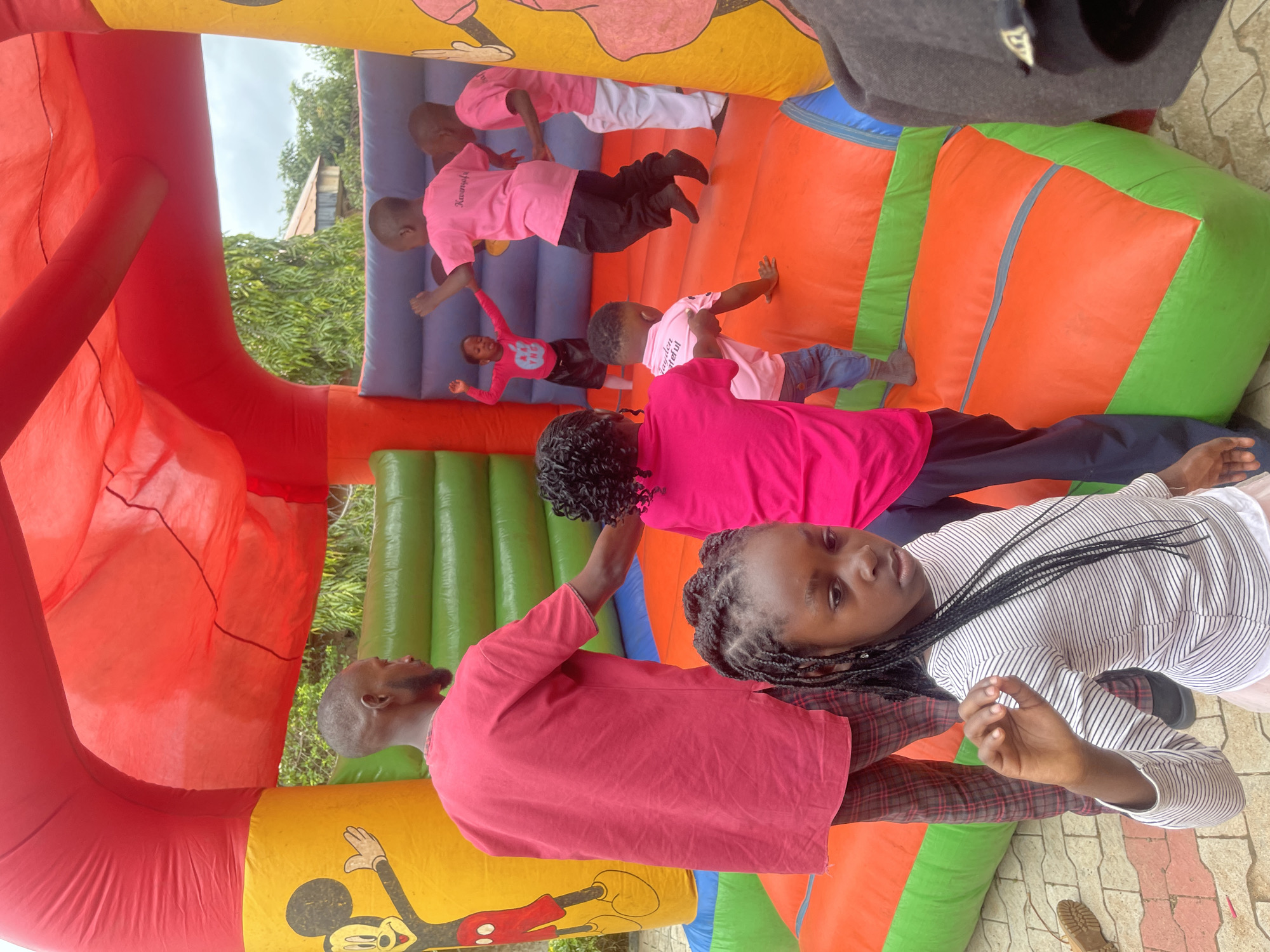 This image captures a vibrant scene at an outdoor bouncy castle event, likely located in Gura Top, Nigeria. The bouncy castle, a dominant feature, is a large, inflatable structure with bright red, yellow, green, and orange panels. Mickey Mouse imagery adorns the yellow sections. Inside the castle, several children are engaged in joyful play. One child in a pink shirt with an apple graphic jumps with excitement, while others in pink t-shirts with text are also actively bouncing. A man in a red t-shirt and plaid pants stands on the edge of the bouncy castle, seemingly supervising or participating. In the foreground, a young girl with braided hair, wearing a striped long-sleeved shirt, looks towards the camera with a thoughtful expression, her hand raised. The ground surrounding the bouncy castle is paved with interlocking bricks. Lush green foliage and trees are visible in the background, suggesting a tropical or sub-tropical environment. The sky appears overcast. The overall atmosphere is one of fun and celebration.