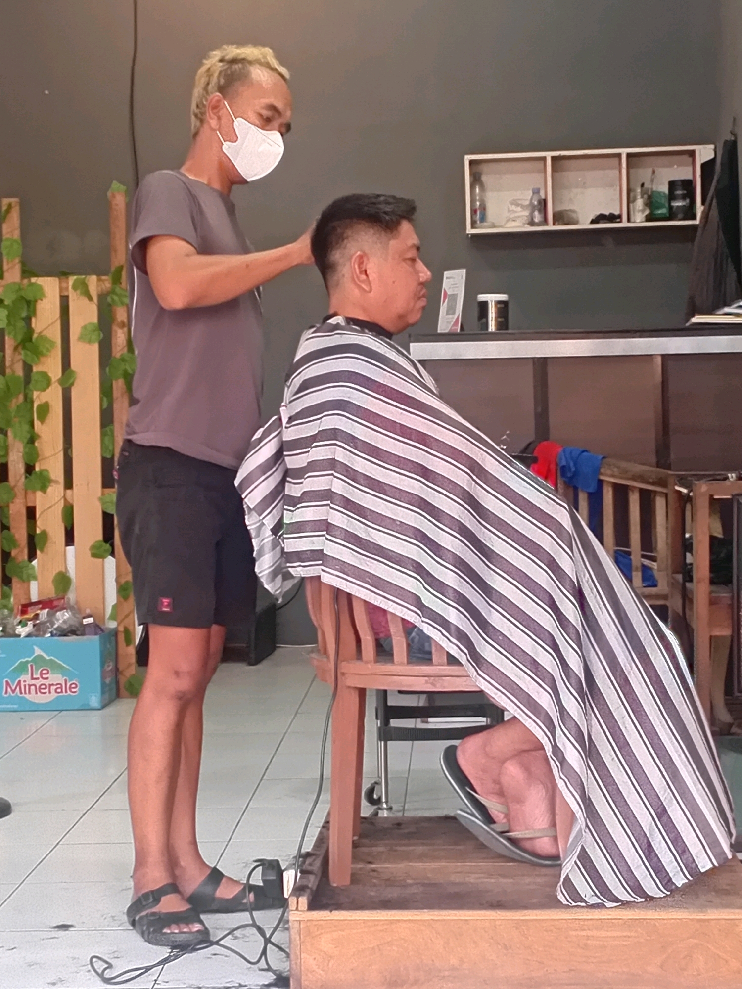 A masked barber with blonde, spiky hair cuts the hair of a man sitting in a chair in what appears to be a small barbershop in Besito, Indonesia. The barber is wearing a grey t-shirt and black shorts. The client is wearing a striped cape. The background shows shelves with bottles and other items, a wooden partition with ivy, and a box of Le Minerale water. The floor is tiled.