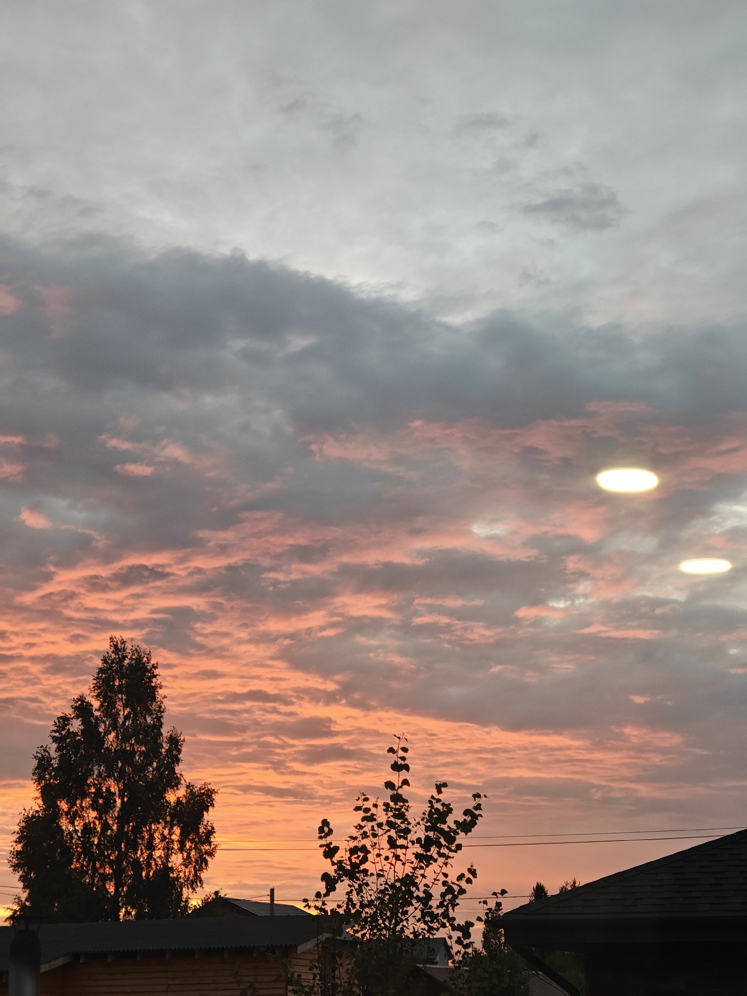 The image depicts a sunset sky over a rural area, likely in Русский Вожой, Russia. The sky is filled with a mix of gray and pinkish-orange clouds, suggesting the sun is low on the horizon. Three bright, circular or oval light sources are visible in the upper right quadrant of the sky, which appear to be artificial lights reflecting off or passing through the clouds. In the foreground, silhouetted against the sky, are two trees, one larger and more prominent on the left, and a smaller one on the right. Below the trees, the dark roofs of several buildings are visible, along with the top portion of a wooden structure. Power lines are also discernible in the middle ground. The scene is devoid of any people or active events.