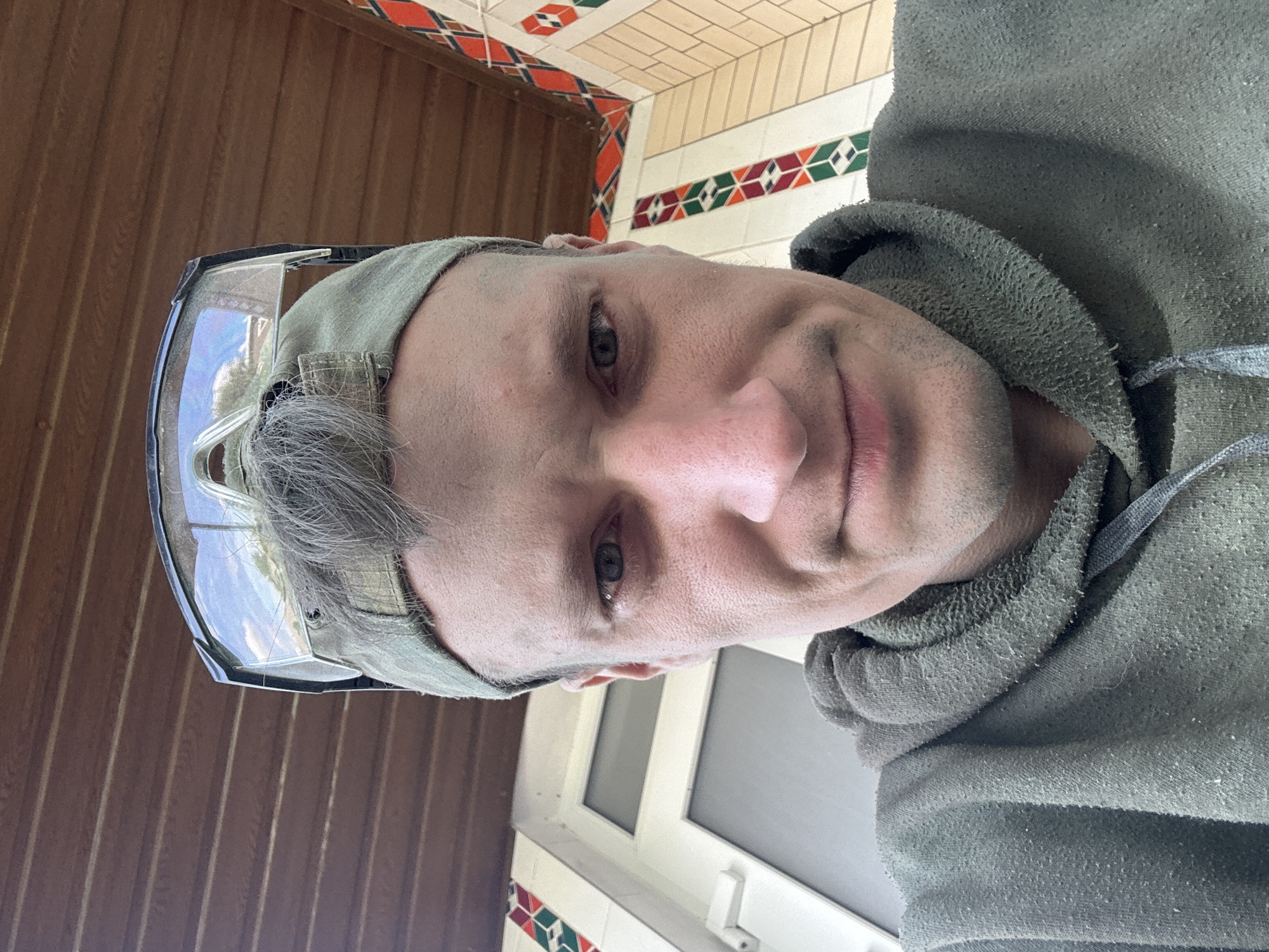 The image is a close-up, low-angle selfie of a man wearing a green hoodie, a green baseball cap, and protective goggles pushed up onto the cap. He has short, light brown hair. His face is looking directly at the camera, with his eyes open and a slight smile. The background shows an overhang with brown wooden planks and a wall with decorative tiles featuring geometric patterns in red, green, and white. To the left of the man, a white door or window frame is visible. The scene appears to be outdoors, possibly on a porch or covered area. There is no discernible indication of the specific location within the city of Makiv, Ukraine.
