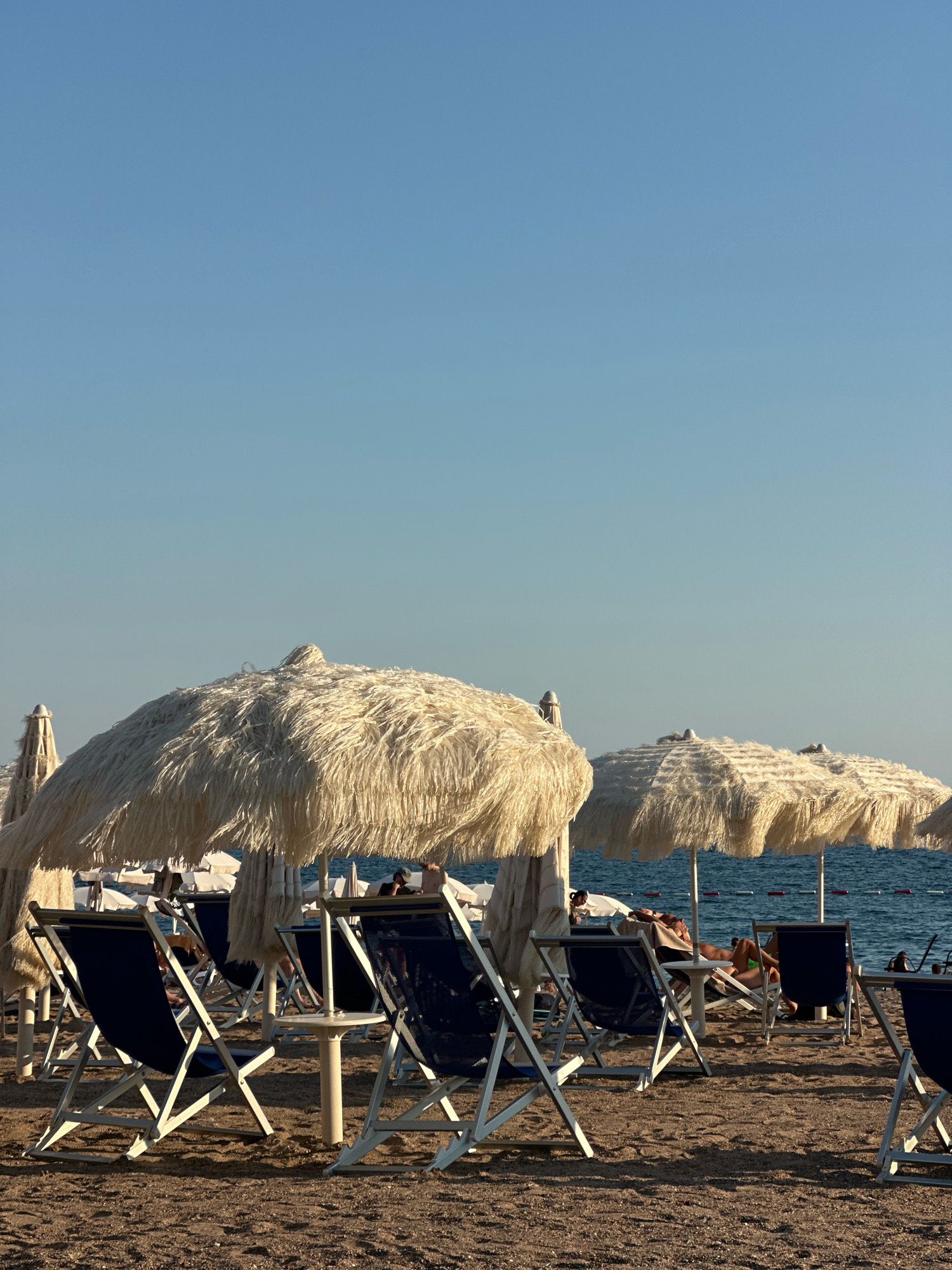 The image depicts a beach scene in Boreti, Montenegro. In the foreground, several empty blue beach chairs are arranged on a sandy beach. Behind the chairs, large thatched umbrellas provide shade. The sea is visible in the background, with a clear blue sky above. A few people are scattered on the beach, some relaxing on loungers under the umbrellas, while others are partially obscured. The lighting suggests it is late afternoon, with the sun casting long shadows.