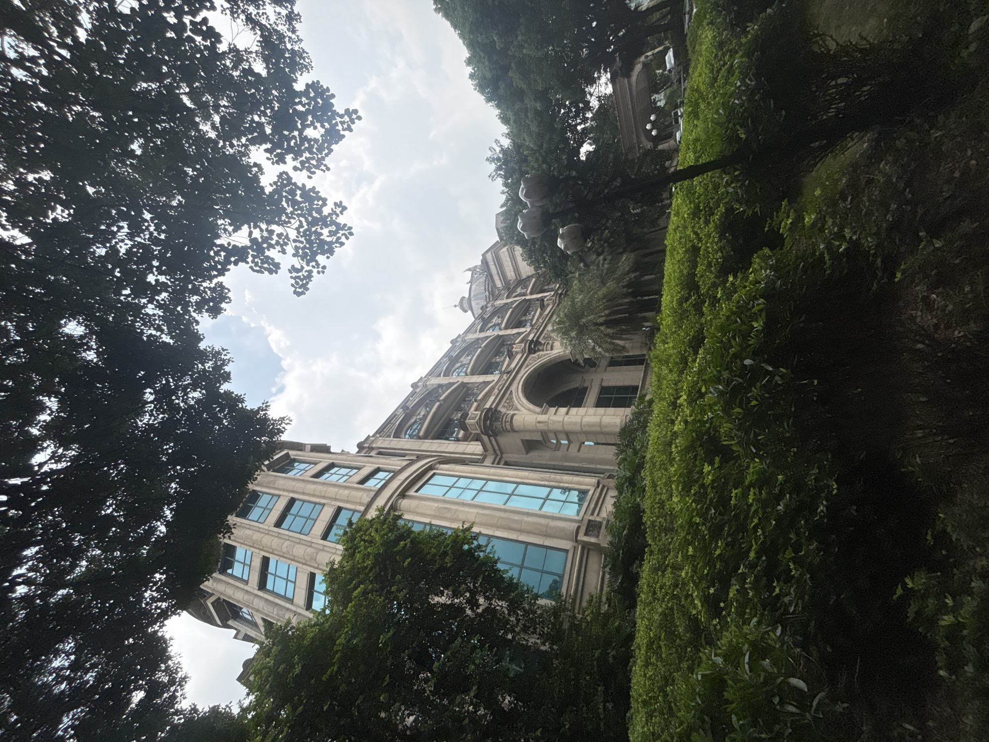 The image depicts a wide-angle, low-angle shot of a grand, multi-story building with a classical architectural style, likely located in Nanhai District, China, given the provided context. The building is constructed of light-colored stone and features numerous large, rectangular windows with a distinctive blue tint. Prominent architectural elements include rounded corners on some sections of the building, arched entrances, and balconies with decorative railings. A large, domed structure is visible atop one part of the building.
The building is surrounded by lush greenery, with dense bushes and trees in the foreground and to the sides. The foliage is a vibrant green, creating a strong contrast with the building's facade. The shot is framed by the dark silhouettes of tree branches and leaves in the upper portion of the image, suggesting the viewpoint is from beneath a canopy of trees. The sky above is partly cloudy, with patches of blue visible. In the background, beyond the immediate greenery, the building extends further, and a covered walkway with arched openings and a street with some vehicles can be glimpsed. A peculiar sculpture resembling large, bulbous shapes, possibly flowers or abstract forms, is situated among the trees to the right of the building's central section. No people are visible in the frame.