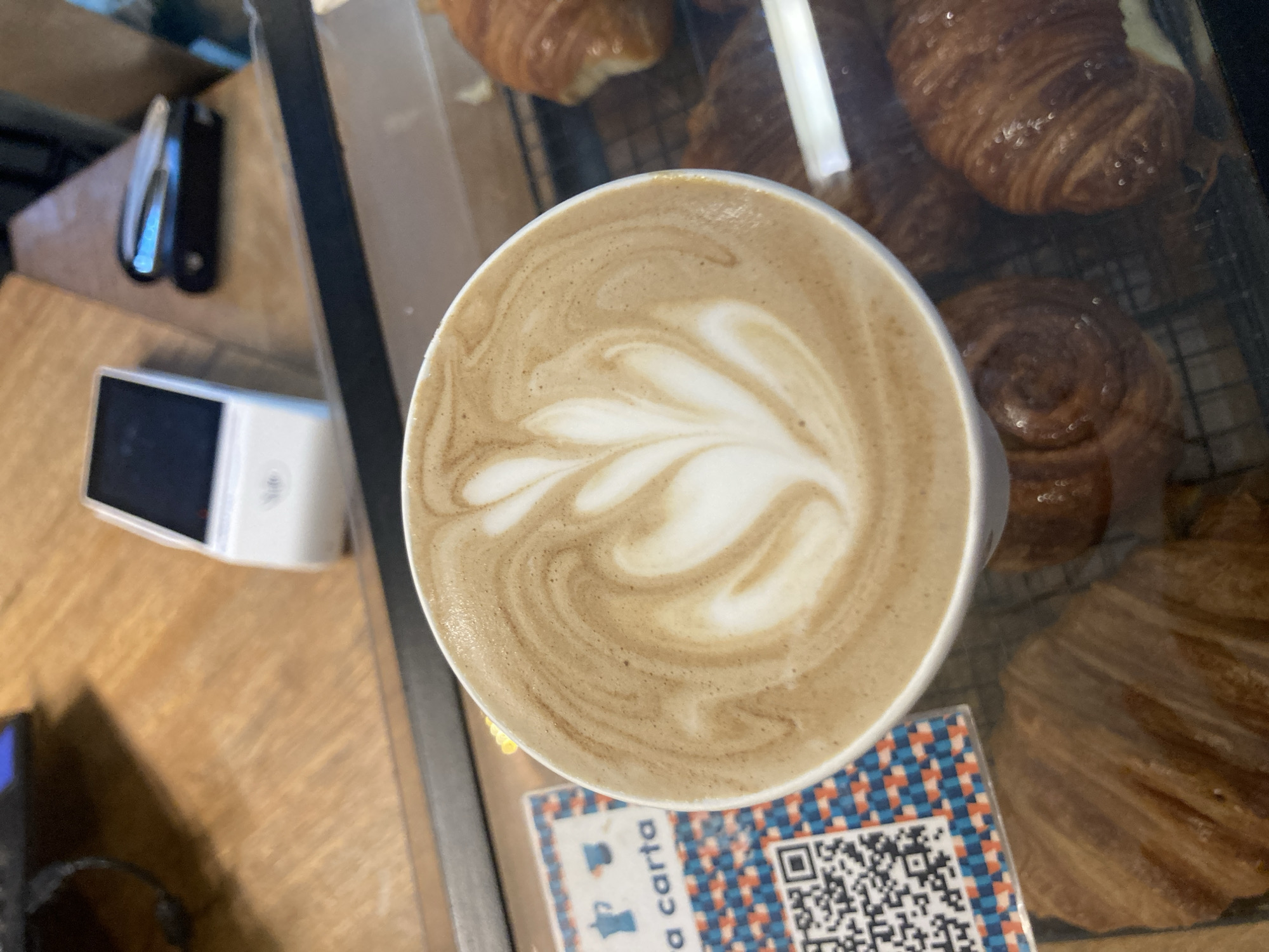 An overhead shot captures a freshly prepared latte with intricate white latte art, resembling a multi-leaf design, centered in a ceramic cup. The frothy beige coffee fills the cup to the brim, featuring swirls and patterns of lighter and darker tones.
The coffee cup is situated on a light wooden counter or table. To the right and in the background, several golden-brown croissants and other pastries are visible inside a clear display case, resting on a metal rack. One large croissant is prominent in the upper right, while a smaller, rolled pastry is seen below it. The reflections on the glass display case suggest an indoor setting, likely a cafe or bakery.
To the left of the coffee cup, partially visible on the wooden surface, is a small, white payment terminal with a dark screen, indicating a commercial establishment. Further left and slightly out of focus, two dark objects, possibly utensils or parts of a coffee machine, are present.
Below the coffee cup, a colorful square card with a blue and red geometric pattern is visible. This card features a QR code on its right side and some text on the left. The visible text reads "a carta," which is Spanish for "menu" or "à la carte," suggesting the location is in a Spanish-speaking region, consistent with the provided context of Buenos Aires, Argentina.
The lighting is bright, illuminating the coffee and pastries, creating an inviting atmosphere often found in a morning or daytime cafe setting.