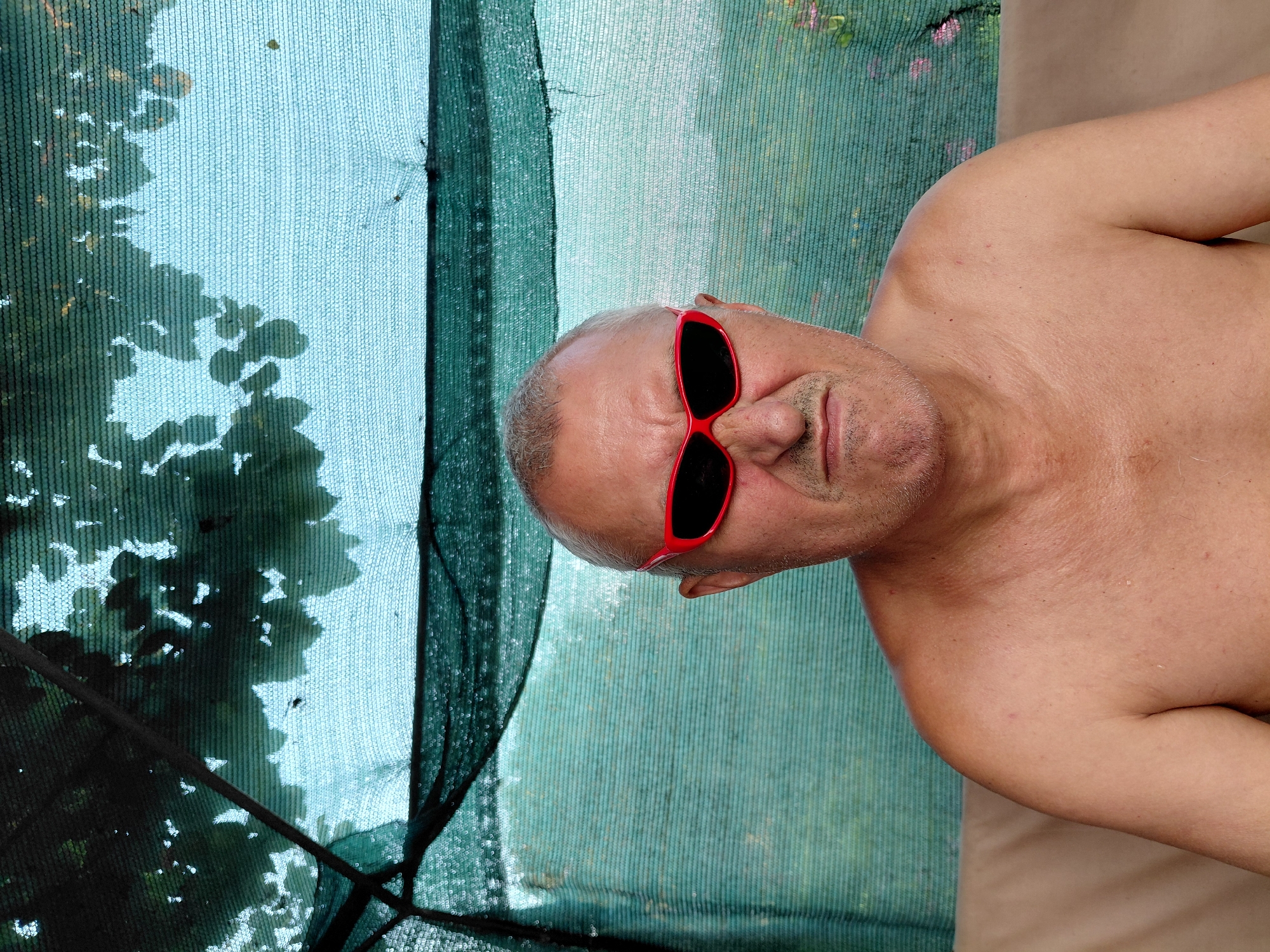 An overhead shot shows a shirtless, middle-aged man with grey hair and a stern expression wearing bright red sunglasses. He is seated on a light brown surface, possibly a sofa, with his bare shoulders and chest visible. The background consists of a green mesh netting, with dappled sunlight and shadows from overhead foliage creating a patterned effect. The overall setting appears to be outdoors, likely in a shaded area like a patio or garden.