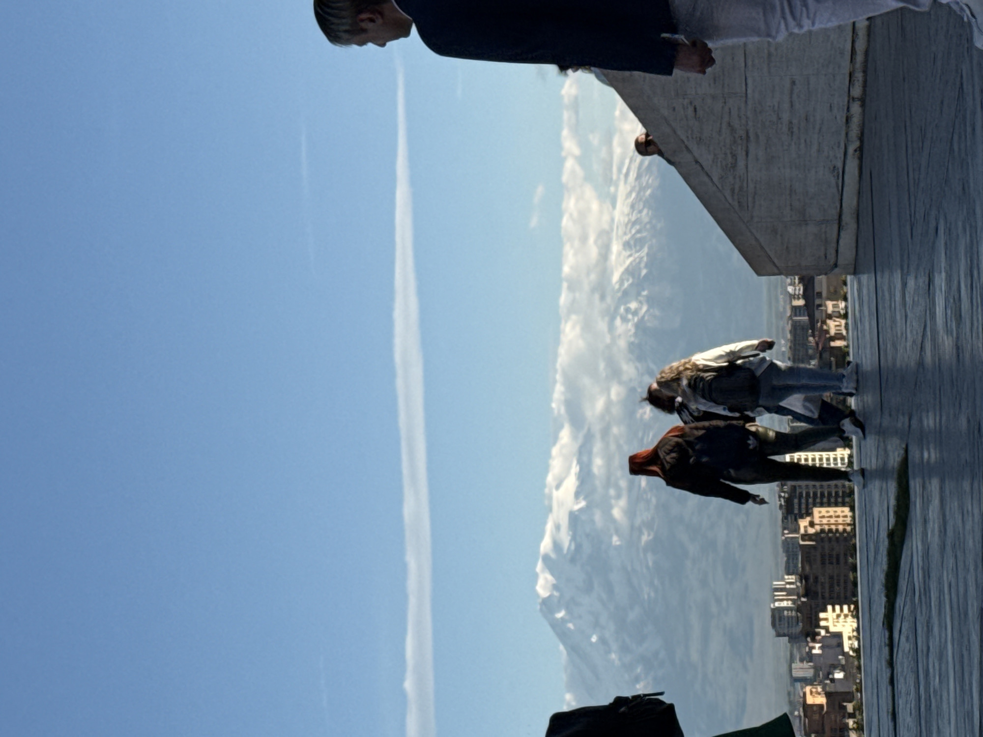 The image depicts a view from a high vantage point in Yerevan, Armenia. A portion of a building's ledge is visible in the upper right corner, with two people standing on it. One person is wearing a navy blue suit jacket and white pants, while the second person's upper body is partially visible. Further down, two more individuals are walking on the ledge. A cityscape is visible below, including buildings and streets. In the background is a large, cloud-covered mountain range. The sky is blue with a single contrail visible. A black bag and a green cloth are in the lower foreground.