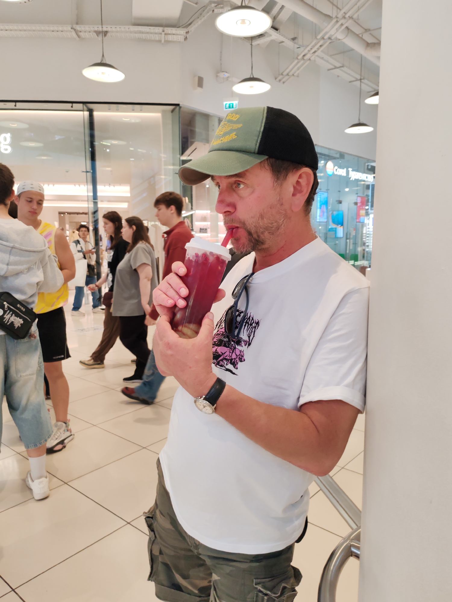 The image shows a man wearing a white t-shirt, camouflage shorts, and a green and black baseball cap, leaning against a white pillar and drinking from a translucent cup with a pink straw. He is wearing a watch on his left wrist. Sunglasses are hanging from the collar of his t-shirt. The background is a retail store with a tiled floor. Several other people are visible in the background, walking in a corridor. The overall environment suggests a commercial setting in a city.